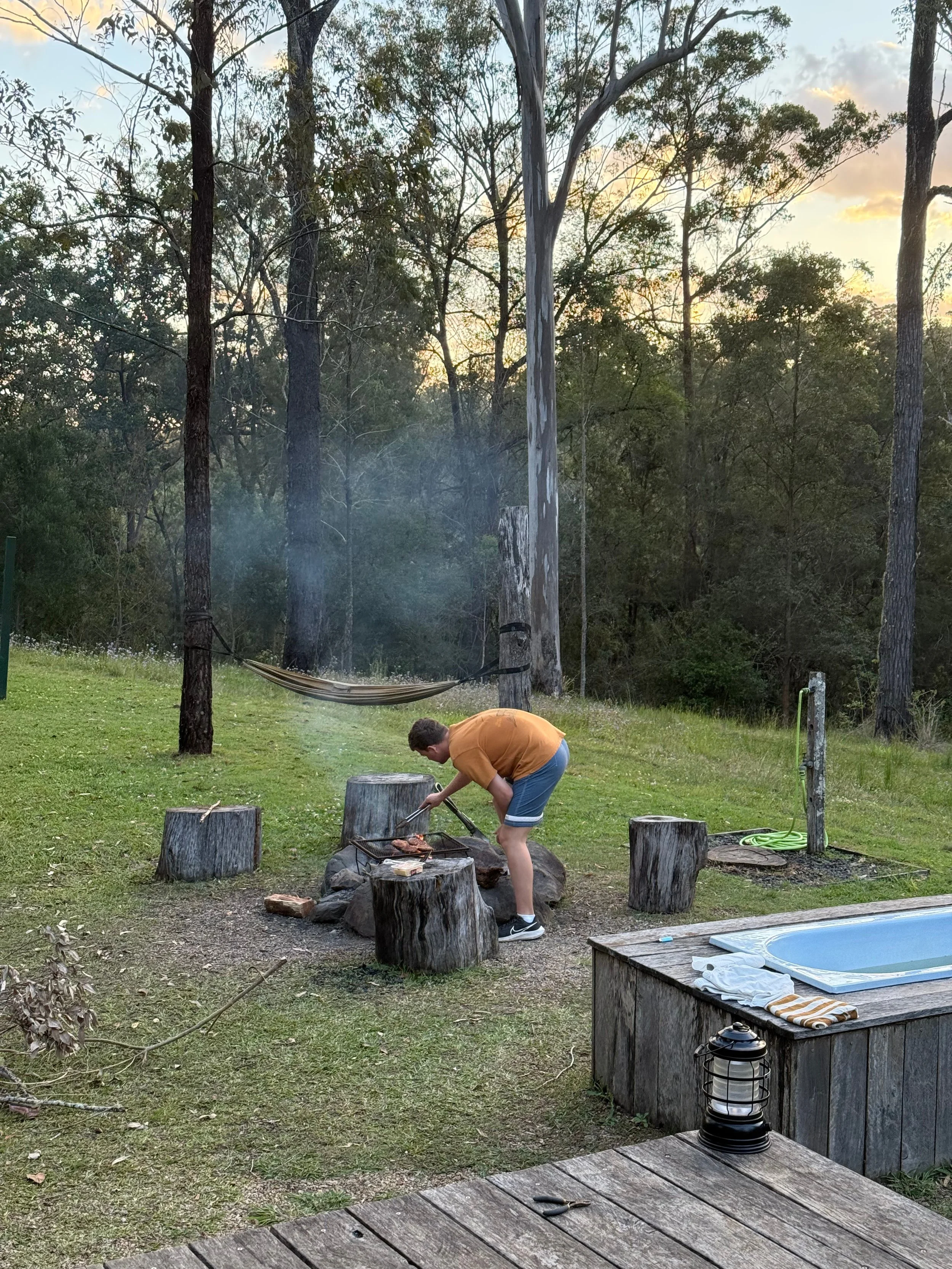 A young boy wearing a mustard yellow shirt and blue shorts is cooking food on an outdoor grill in a backyard near several tree stumps. The backyard has a grassy area, a hammock hanging between trees, and a wooded area in the background. The sky is partly cloudy with the sun setting.