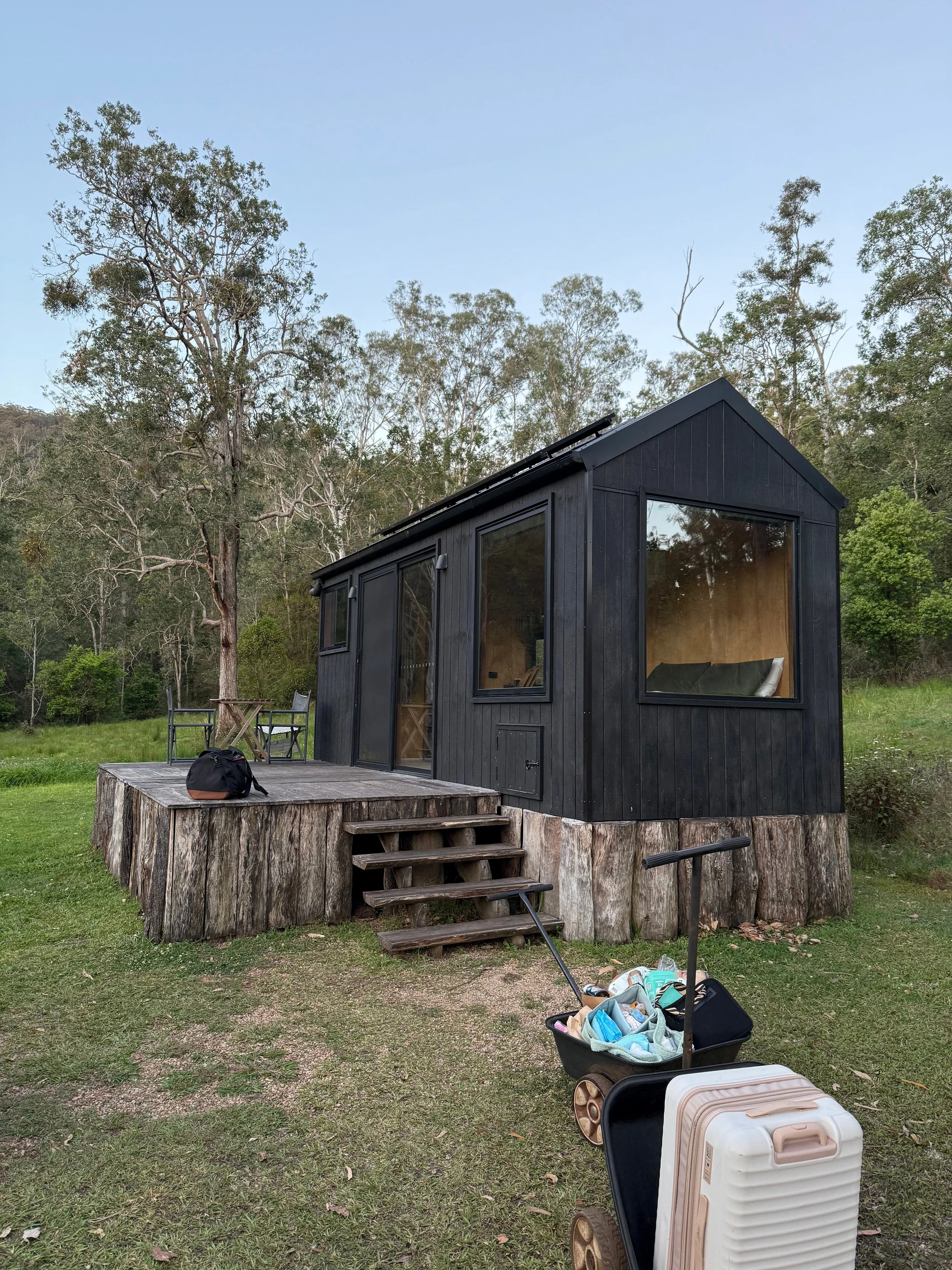 A black tiny house or cabin with large glass windows on a forested hill, surrounded by trees and grass. There is a small wooden deck with chairs and stairs, as well as luggage and a bag nearby.