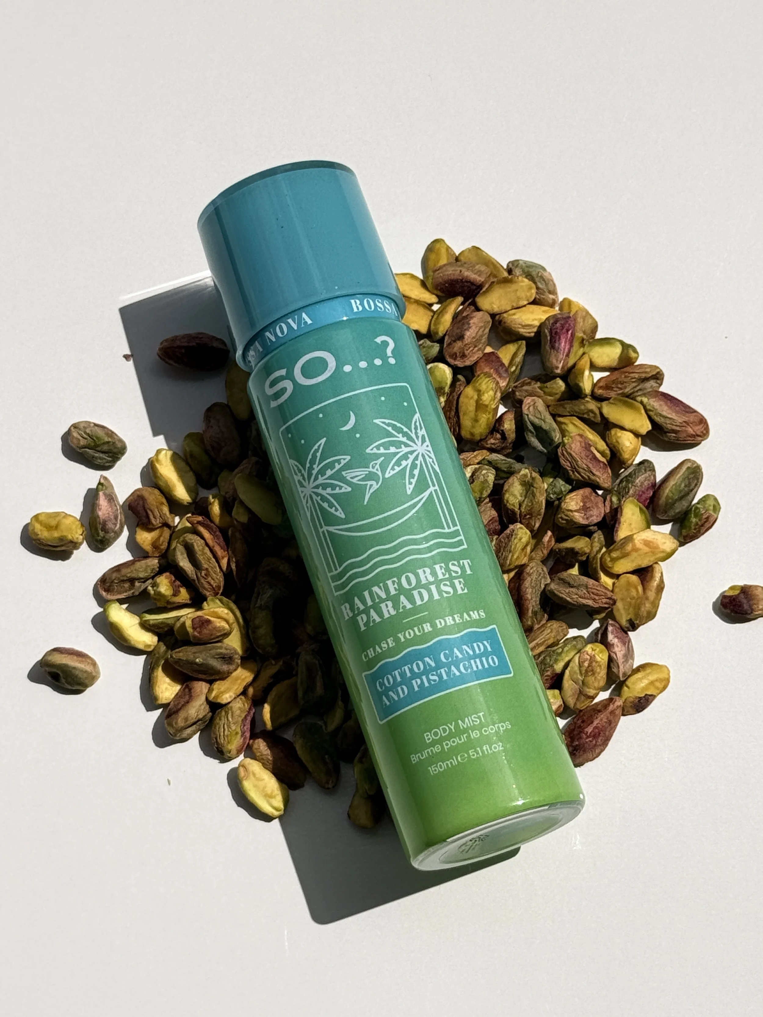 A body mist spray bottle labeled 'Rainforest Paradise' surrounded by scattered pistachio nuts on a white background.