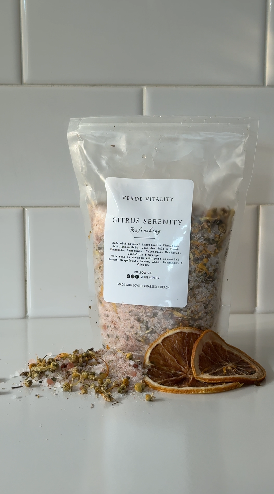 A clear plastic bag of Himalayan bath salts labeled 'Verde Vitality Citrus Serenity Refreshing', with dried orange slices and loose bath salts with herbs and flowers arranged in front.