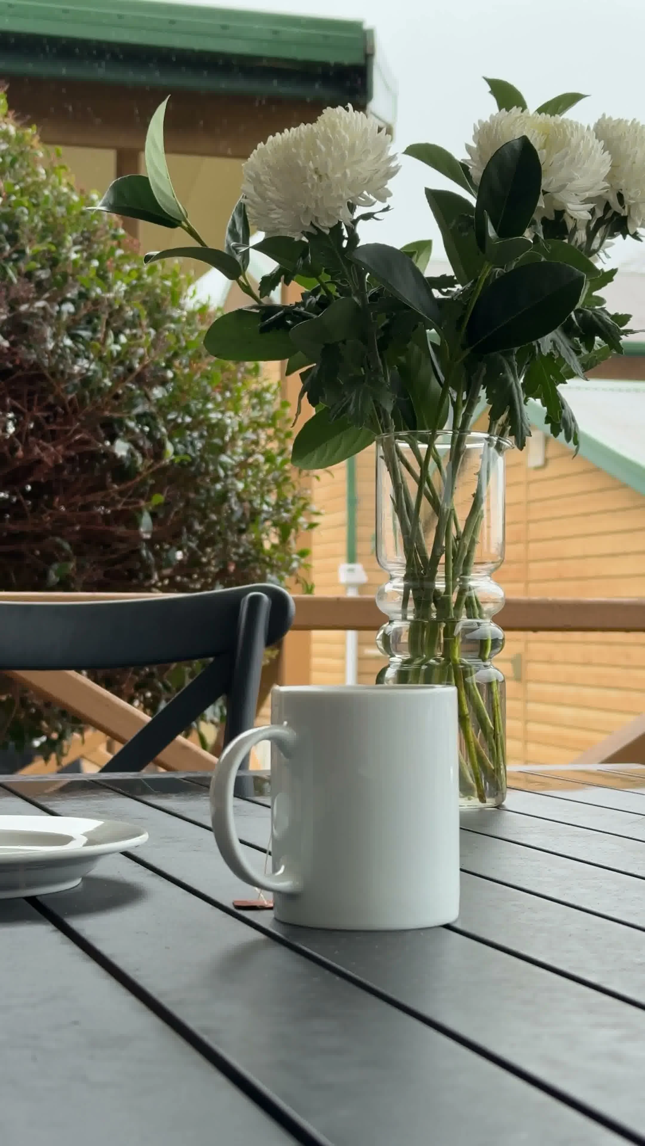 A white mug with a handle sitting on a gray table with wooden planks, in front of a glass vase holding white flowers and green leaves, with outdoor furniture and a wooden fence in the background.