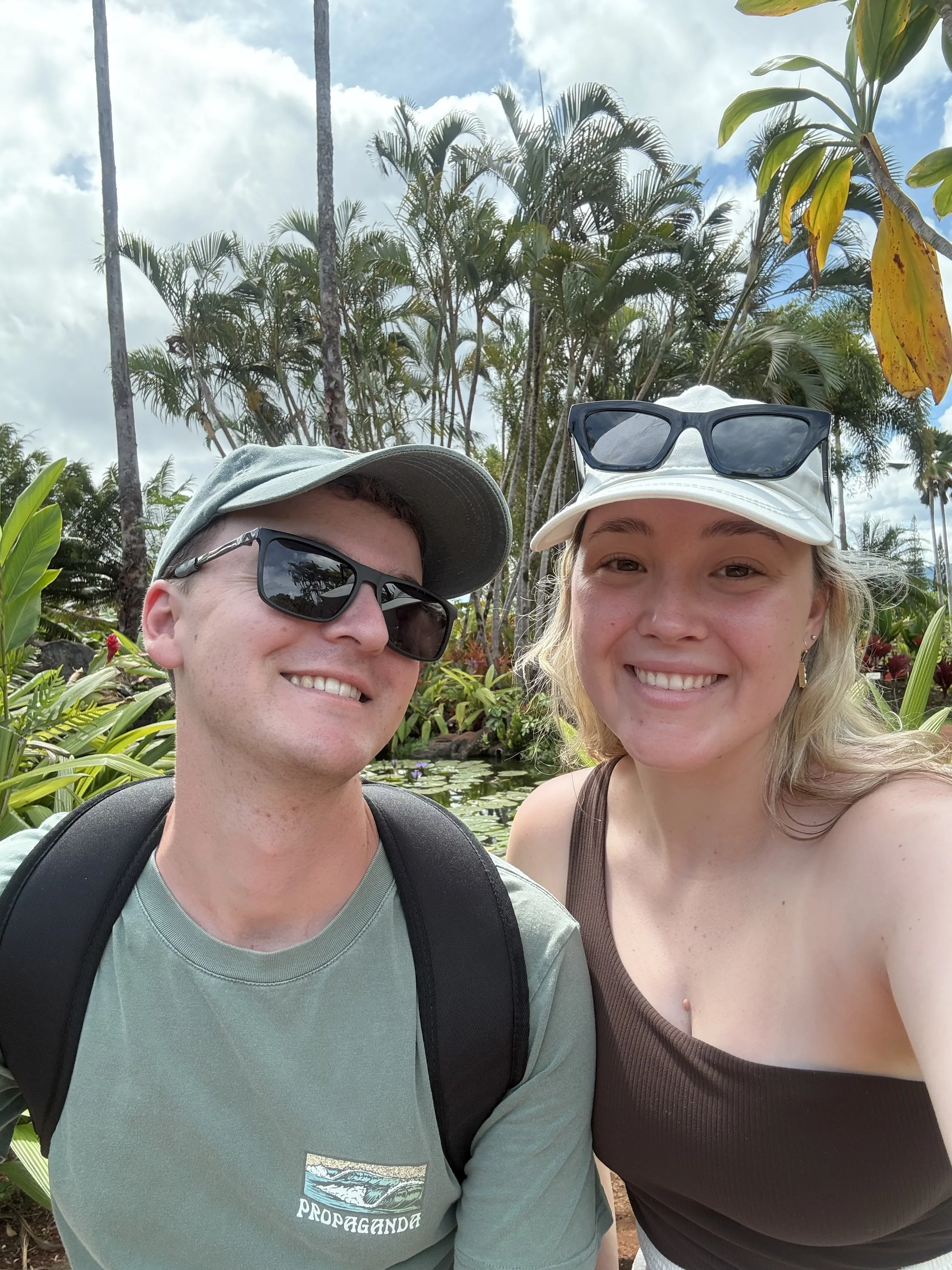 Two people taking a selfie outdoors, surrounded by tropical plants and trees, with partly cloudy sky in the background. The man on the left is wearing sunglasses, a gray cap, a gray t-shirt, and has a backpack. The woman on the right is smiling, wearing a white cap with sunglasses on top, and a brown tank top.