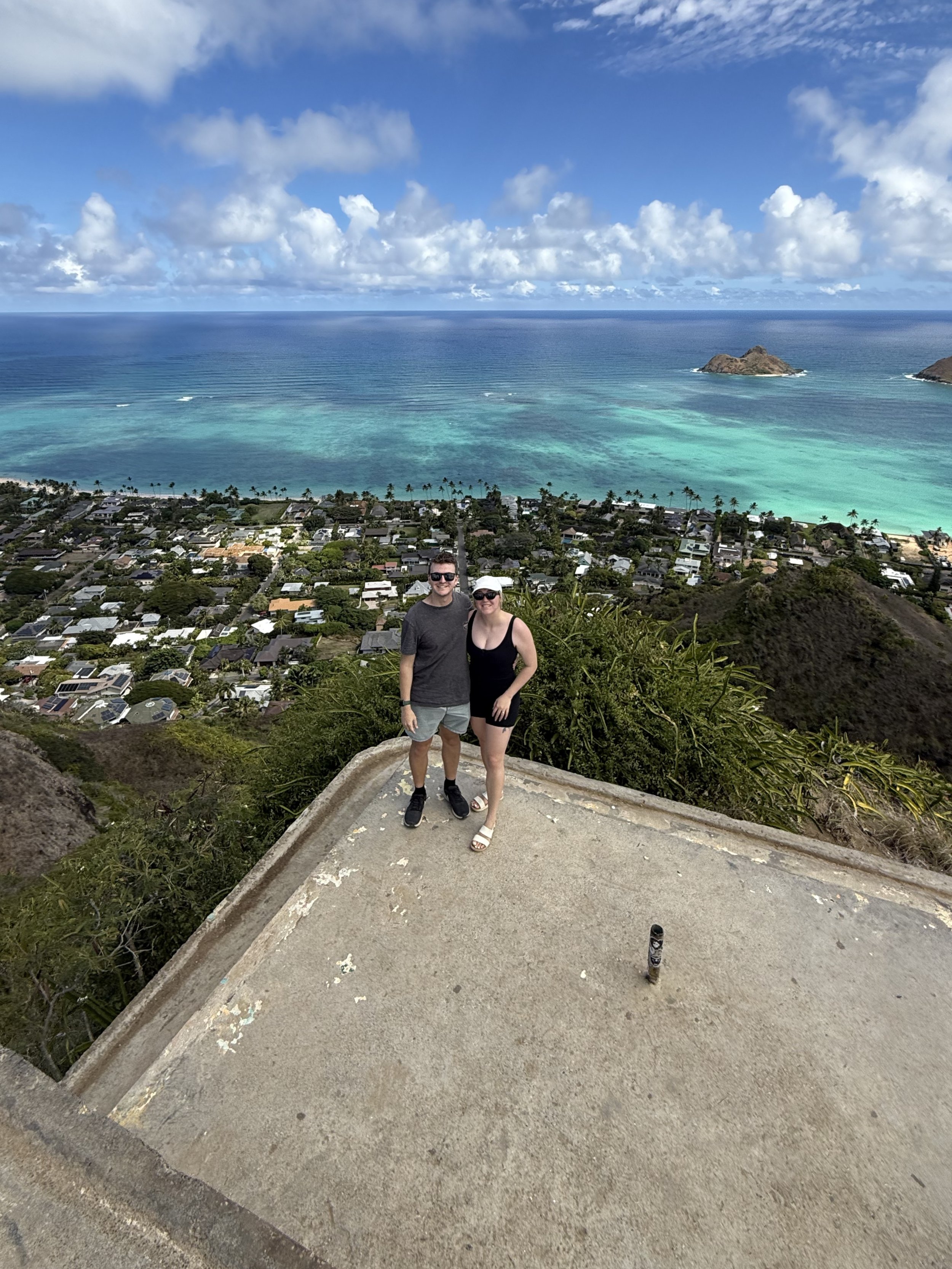 A couple standing on a viewpoint with a coastal town, ocean, and small islands in the background.