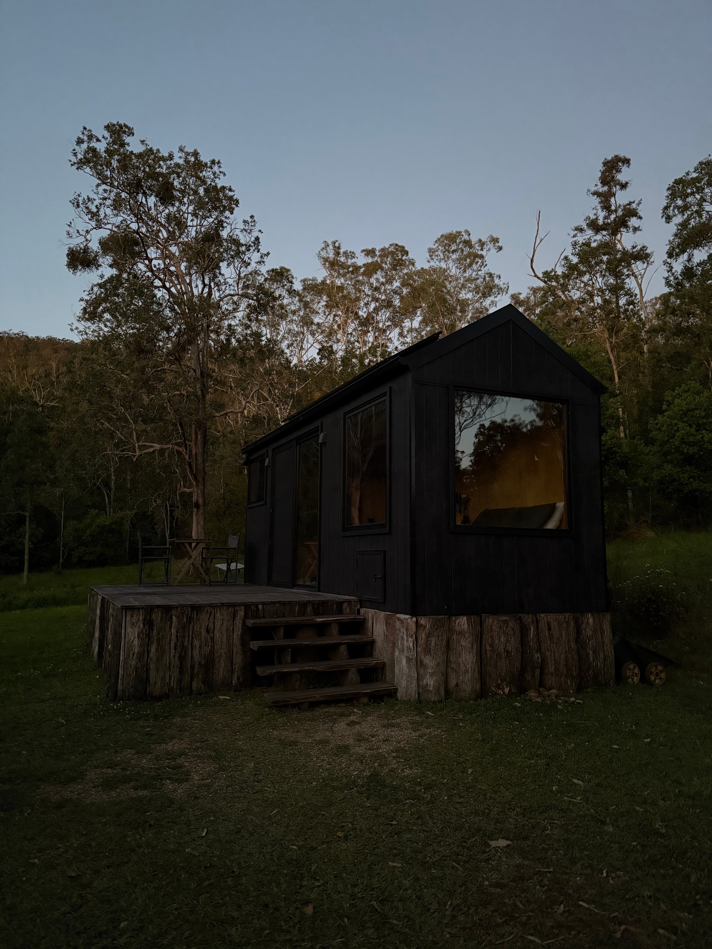 A small black cabin with large windows, built on a wooden platform with stairs, set in a wooded area during dusk or early evening.