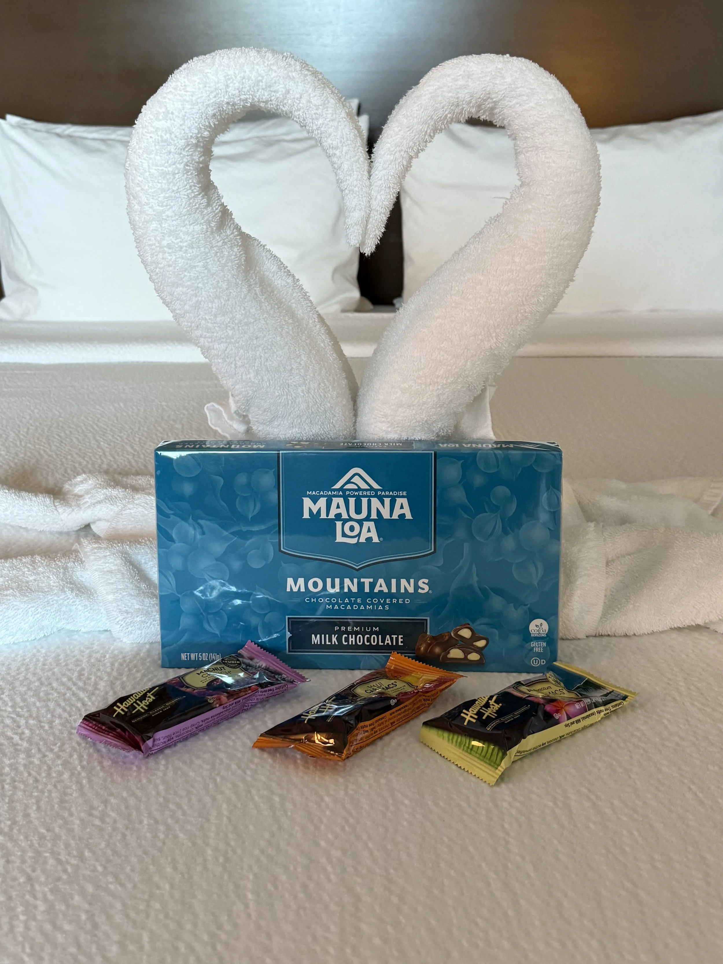 A blue box of Mauna Loa Mountains chocolate-covered macadamia nuts in front of a towel folded into two towel swans with heart shapes, on a bed with white sheets and pillowcases.