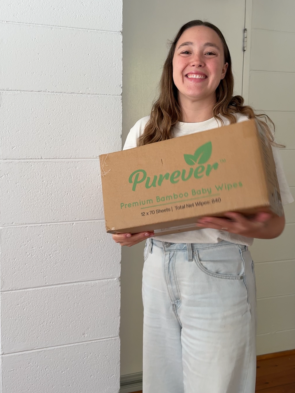 A young woman with long brown hair, smiling, holding a box of Purever bamboo baby wipes in front of a white wall.