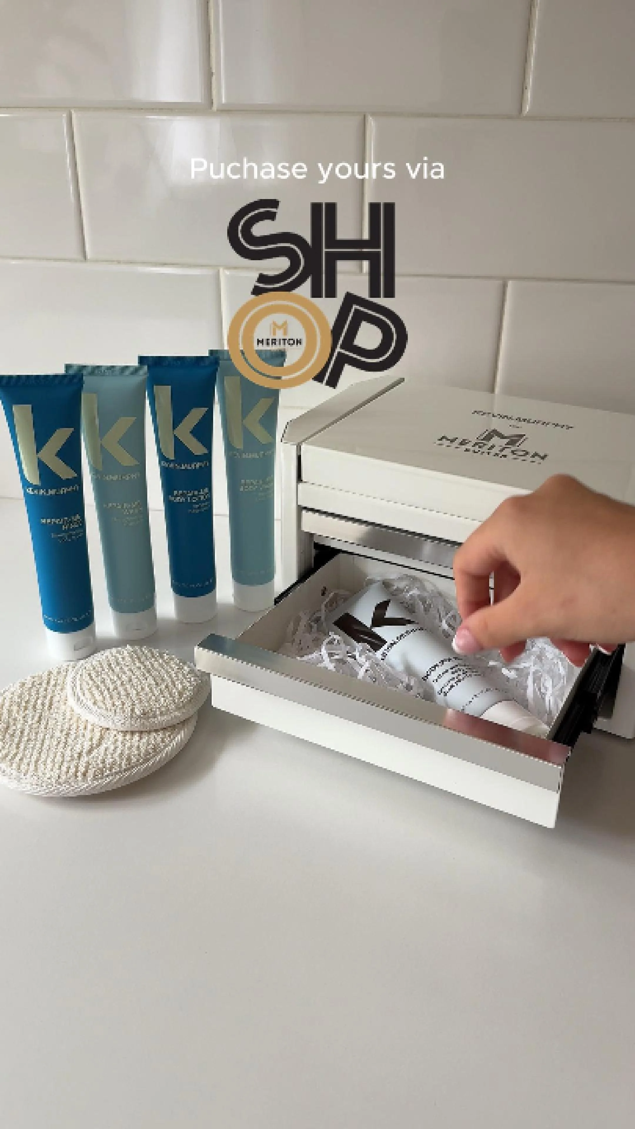 A person opening a box with skincare products inside, four blue tubes labeled 'K' lined up on a white surface, beige textured items, and a white tiled wall in the background with an overlay text 'Purchase yours via SHOP'.