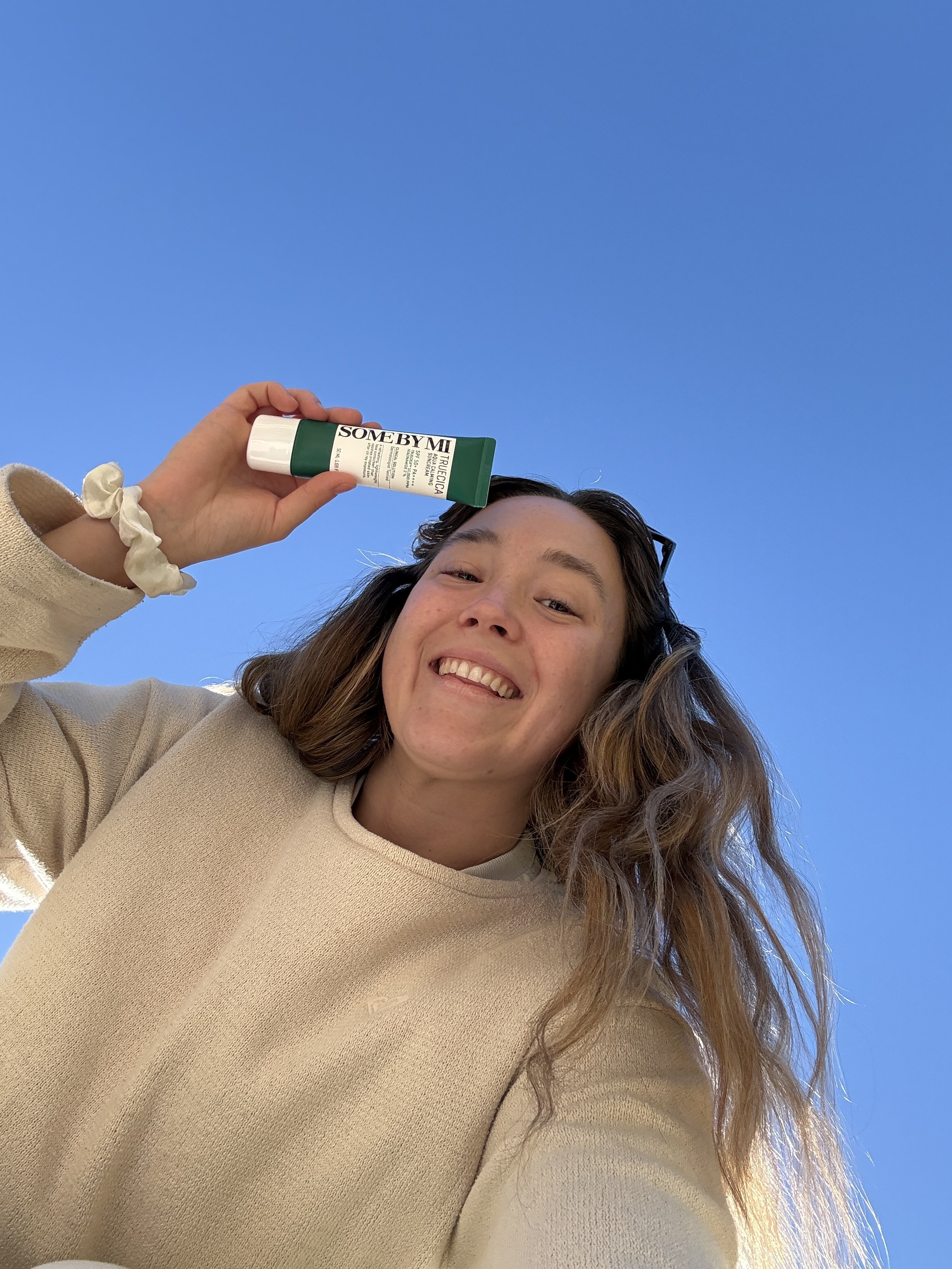 A woman with long wavy hair smiling and holding a tube of SO ME BY MI skincare product against a clear blue sky.