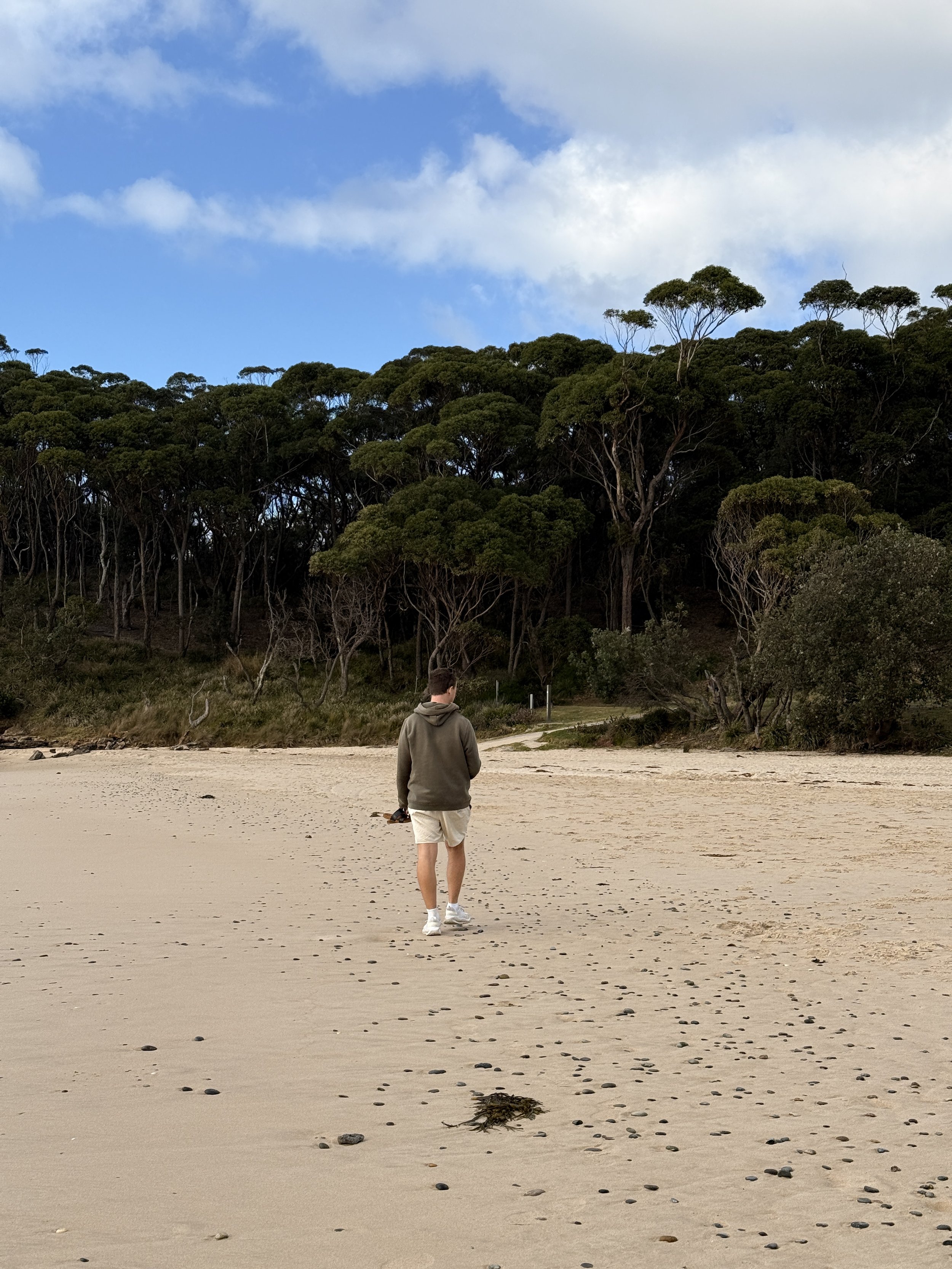 A person in a gray hoodie and white shorts walking on a sandy beach with small rocks, near a forested area and a partly cloudy sky.