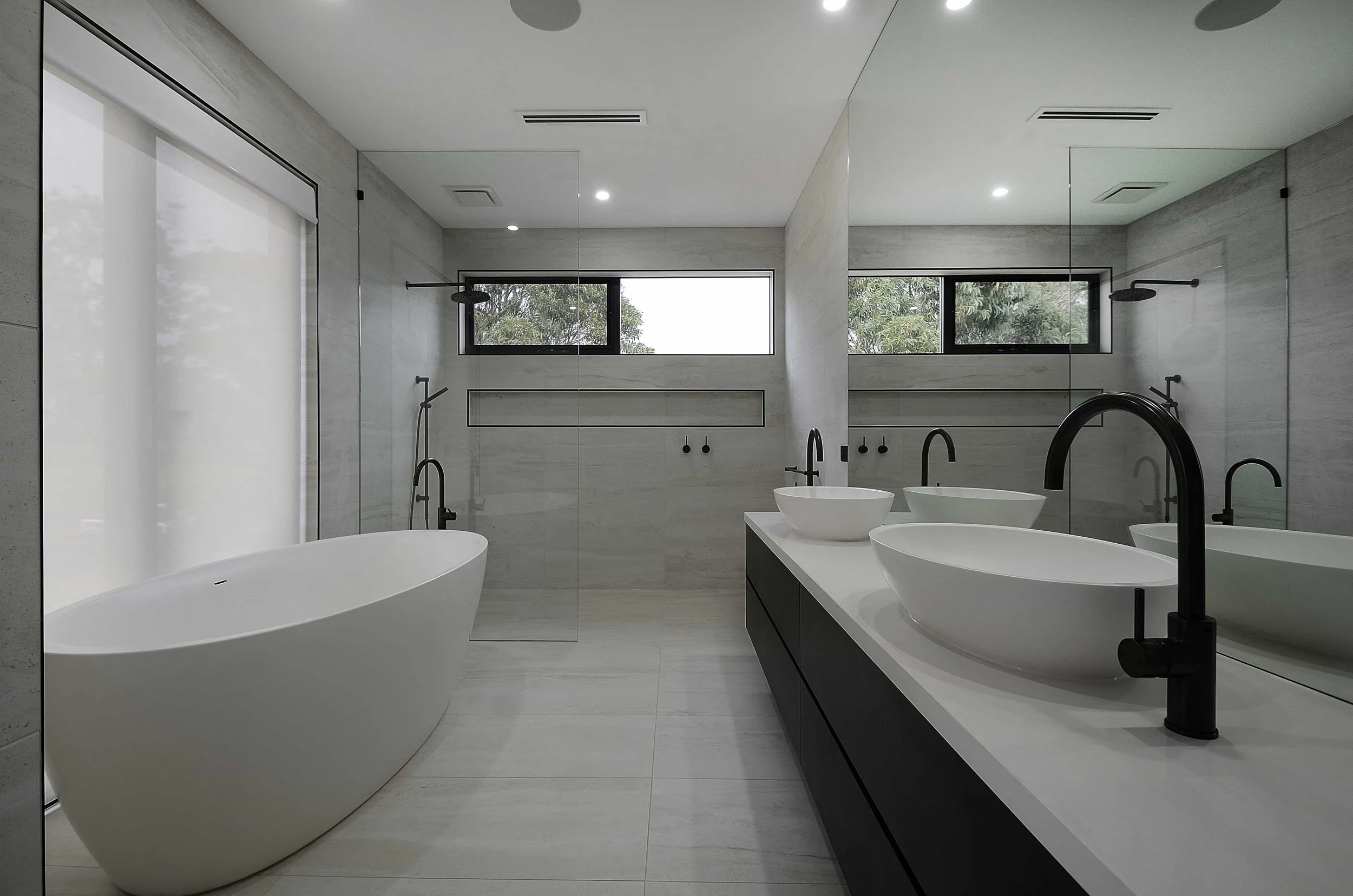 A modern ensuite with a freestanding bathtub, double basins with black faucets, a large mirrored splashback behind the vanity, and a half enclosed walk in shower with a tiled niche on the wall.