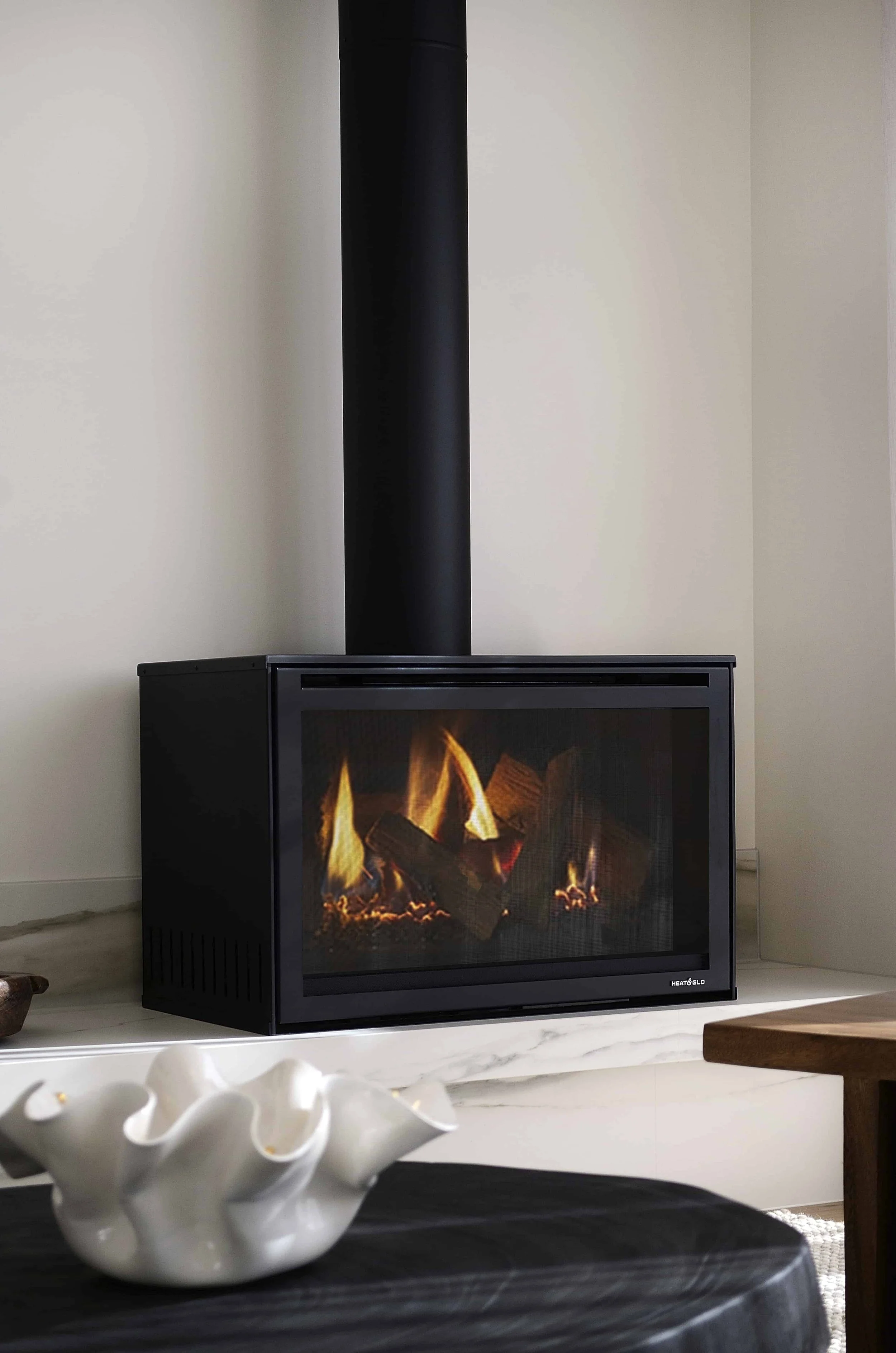 A black fireplace with a glass front showing a cozy fire inside, placed on a white stone benchtop. In the foreground, there is a white ceramic decorative bowl with an abstract, wavy design. The background shows off white walls.