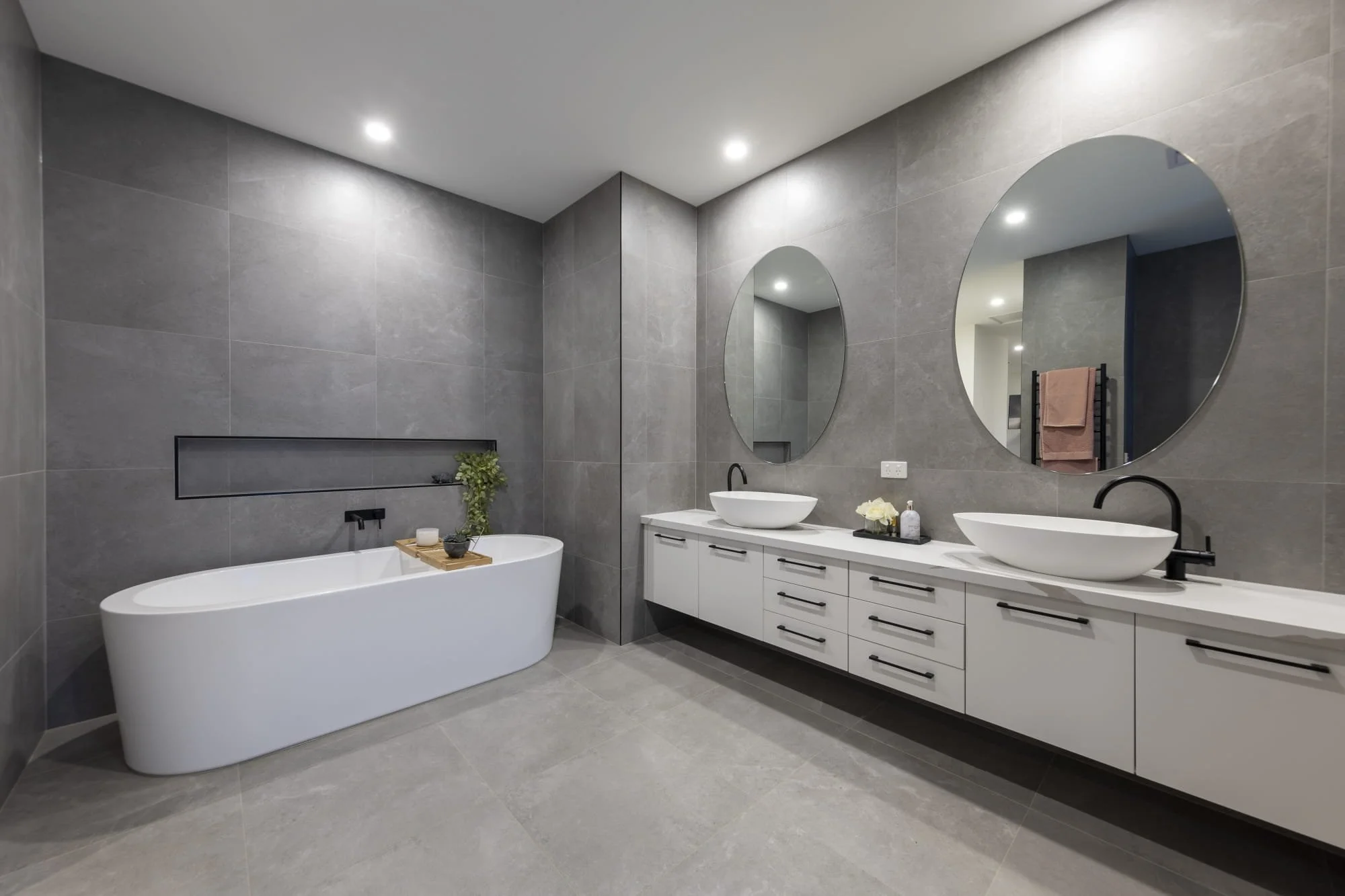 A modern ensuite with grey tiled walls and floor, white vanity with double basins, large oval mirrors, black faucets, freestanding bathtub with a wooden tray and plants on it, recessed niche with decor, and ceiling downlights.
