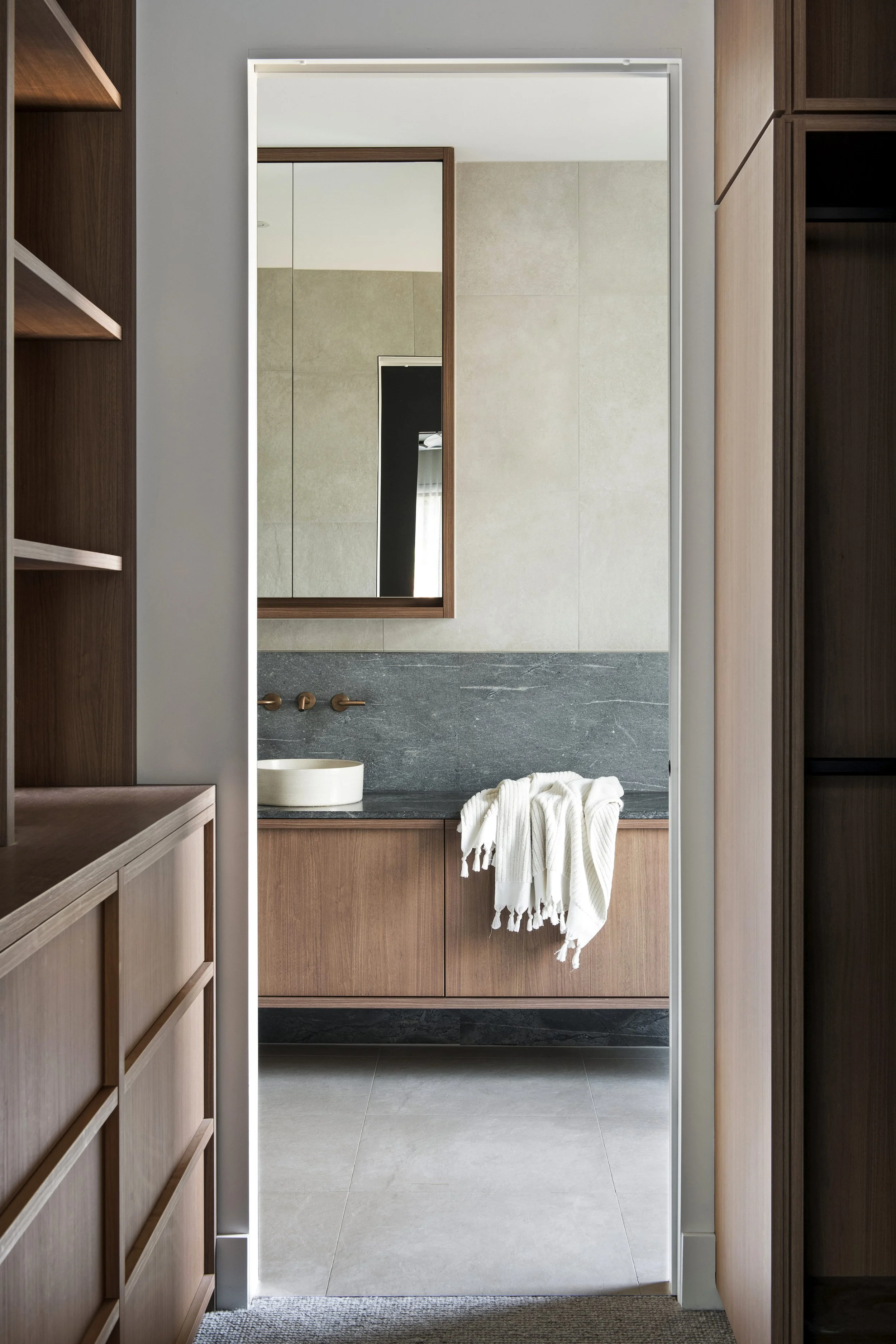 A bathroom seen through a doorway with built in timber cabinetry on both sides, a vanity with a dark stone benchtop, a round above bench basin, and a large mirror with a wooden frame reflecting a black door and a window with curtains.