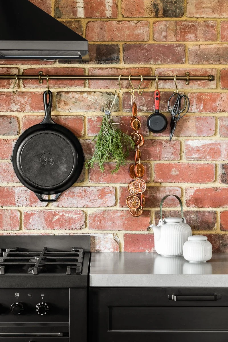 A kitchen with a brick wall, a black cast iron skillet hanging from a hook, dried herbs, dried citrus slices, a small frying pan, kitchen scissors, and a white teapot with a matching smaller container on the benchtop.