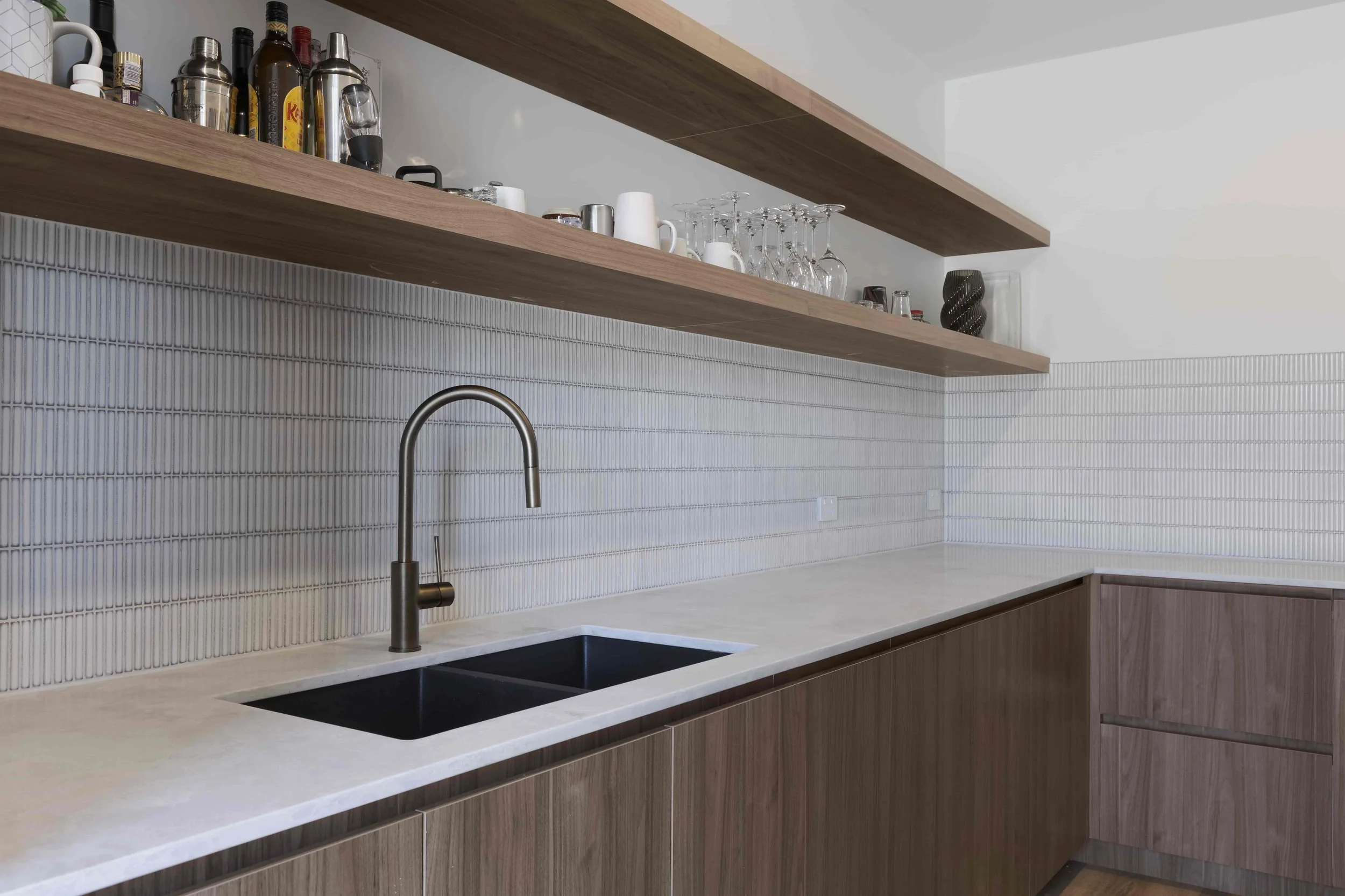 Modern butler's kitchen with a white stone benchtop, a black sink, a gunmetal grey faucet, and two floating timber shelves holding drinking glasses, mugs, and bottles against a white finger tiled wall.