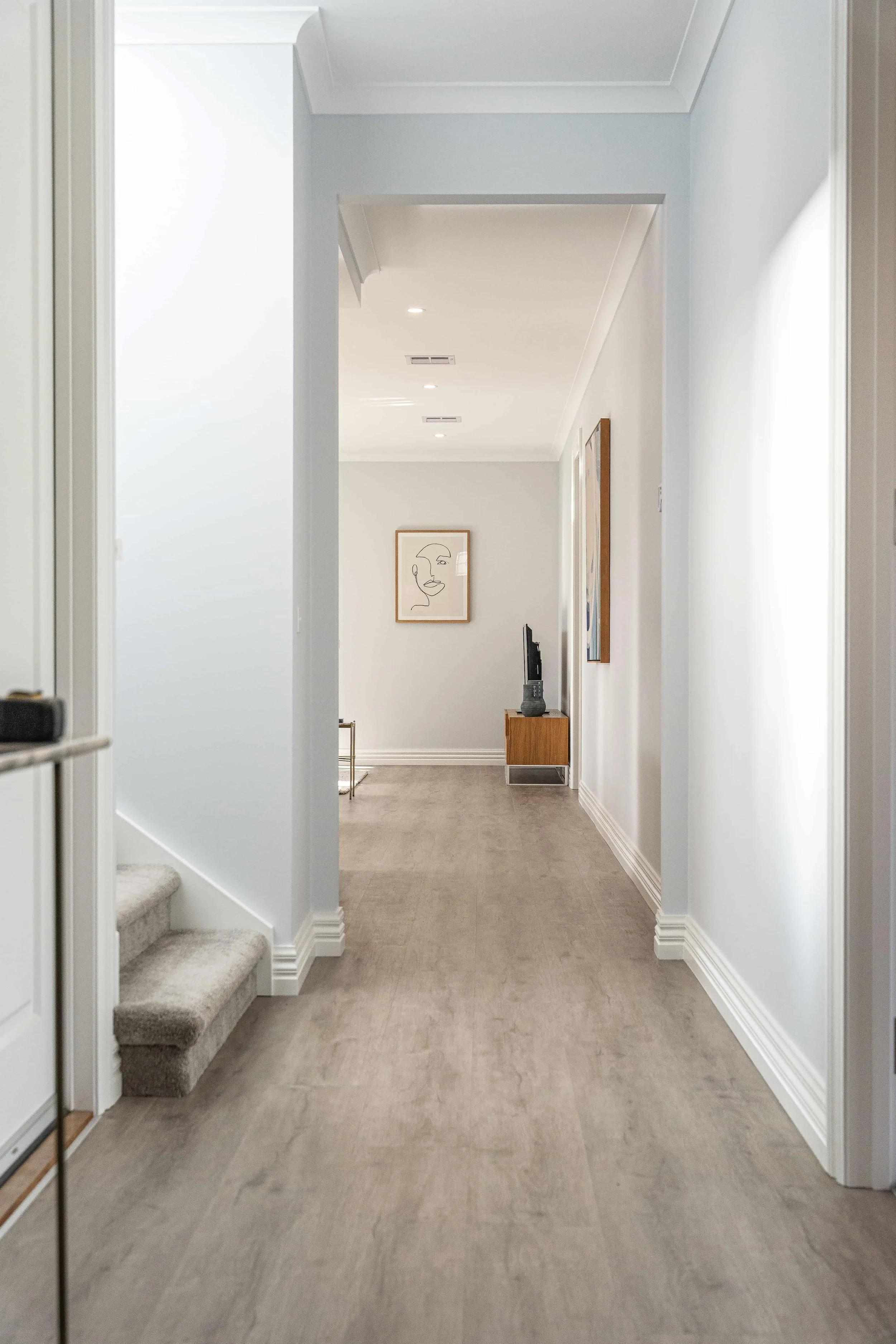 A hallway in a house with light-colored walls and carpeted stairs on the left leading upwards, and framed artwork on the walls. The hallway has a wooden cabinet with a small TV on top and a framed picture nearby.