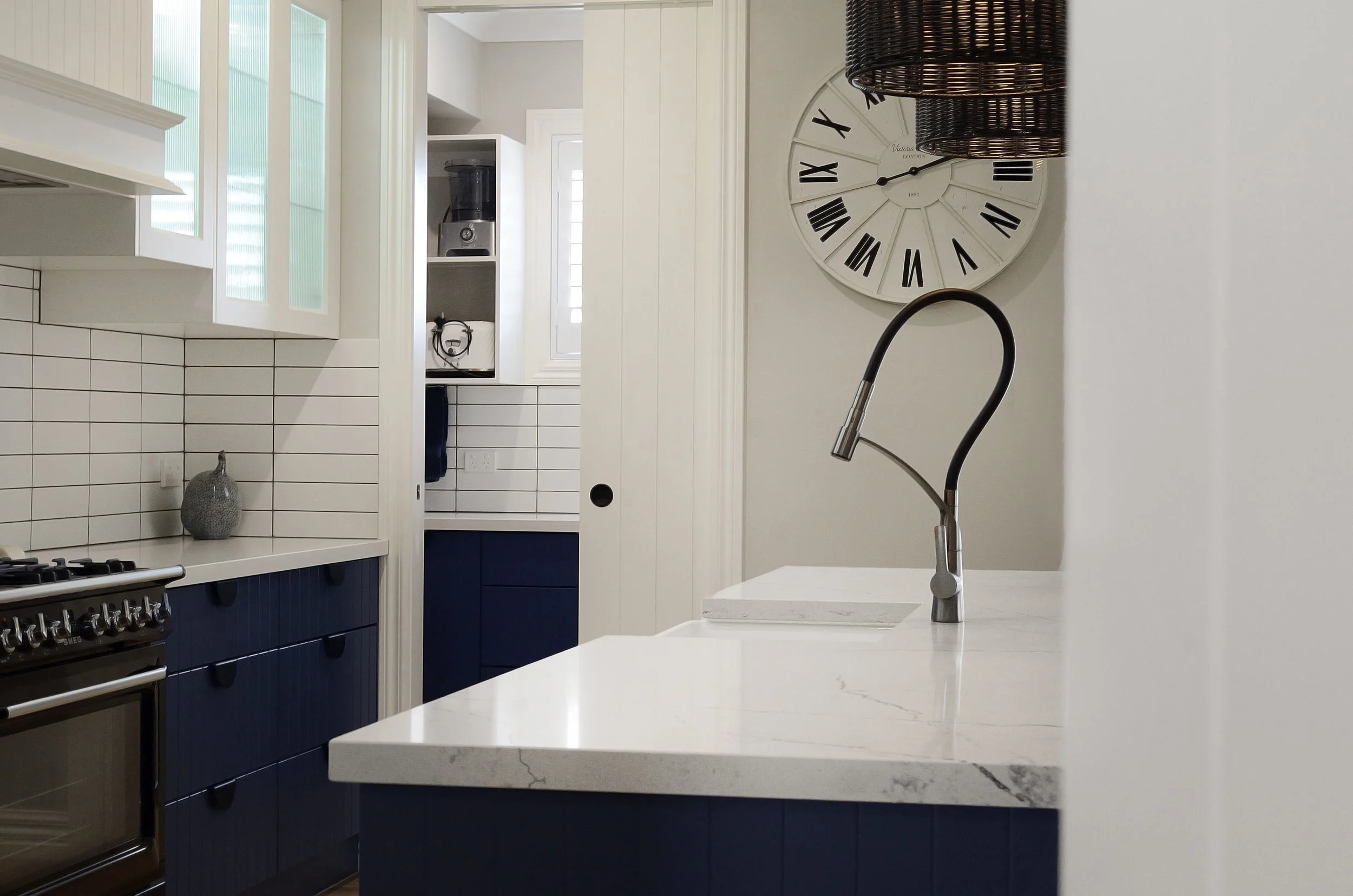 Modern kitchen with white stone benchtop, dark blue cabinets, a white subway tiled splashback, and a large wall clock with Roman numerals. A stainless steel faucet is visible in the foreground.