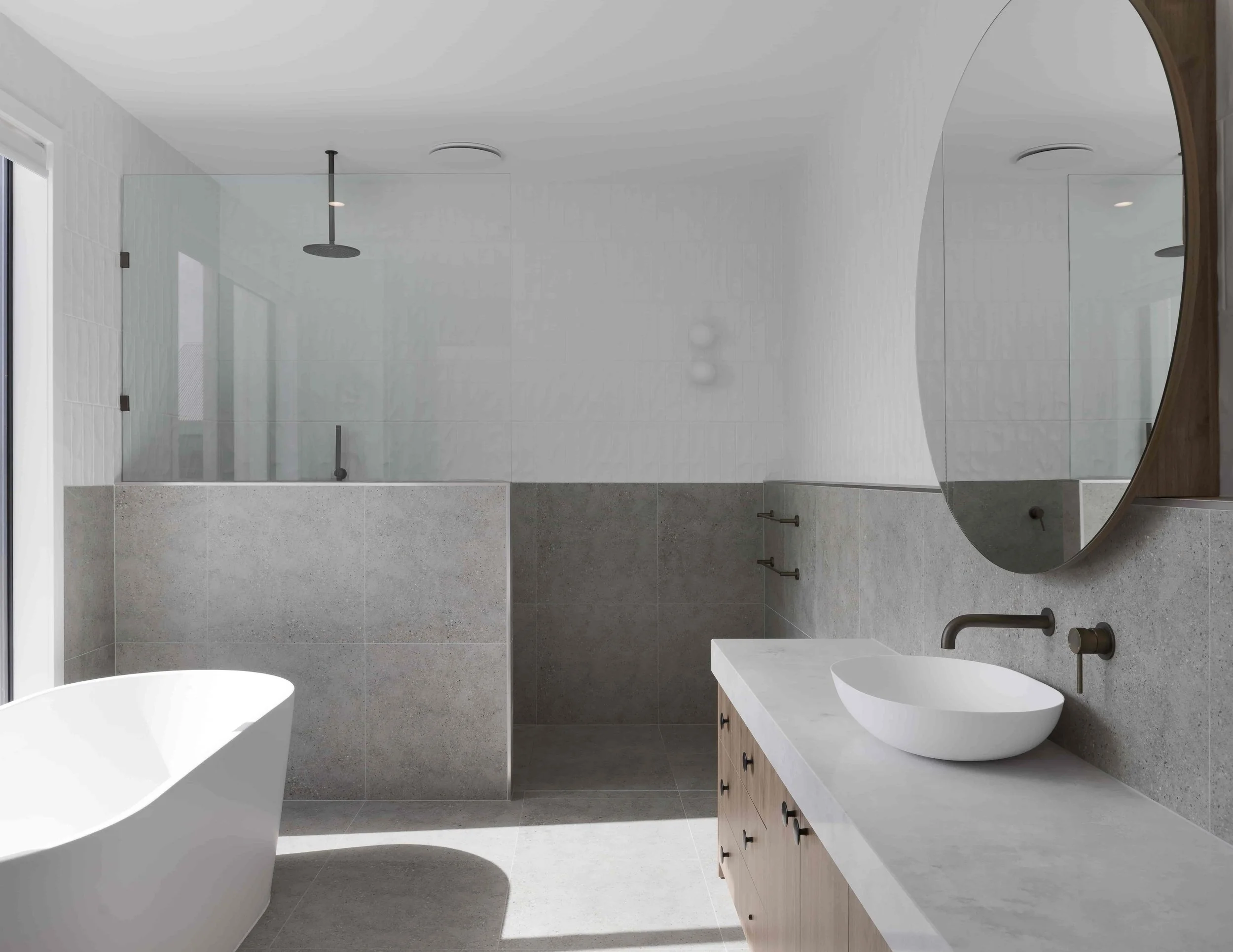 Modern bathroom featuring an open walk-in shower with glass enclosure, rain shower head, white freestanding bathtub, white basin and grey stone benchtop on a wooden vanity, round mirror, and grey tiles on the walls and floor.