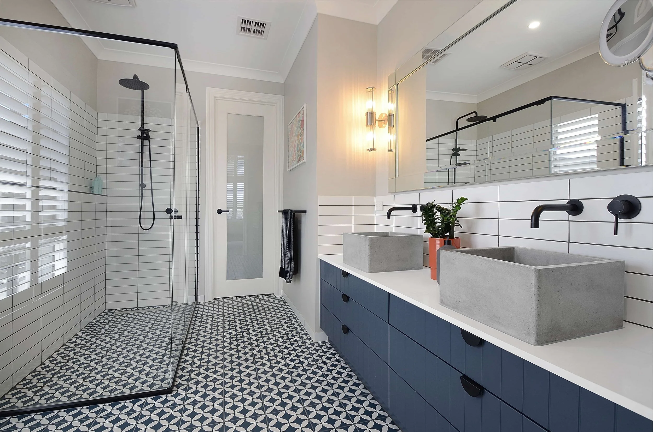 Modern ensuite with a patterned navy and white floor, white subway tiled walls, a large mirror, two concrete sinks with black fixtures, a navy blue vanity, and a glass enclosed shower area with black shower fixtures.
