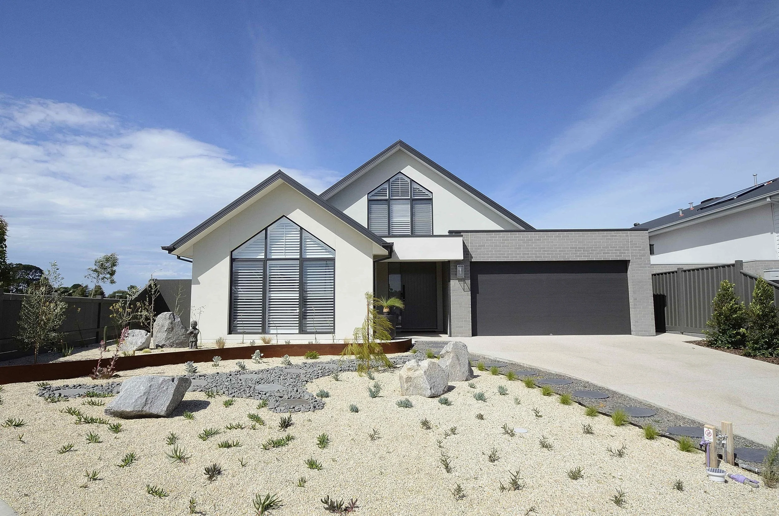 Modern house with large windows, gable roofs, a grey garage door, landscaped front yard with rocks, small plants, and a clear blue sky. 