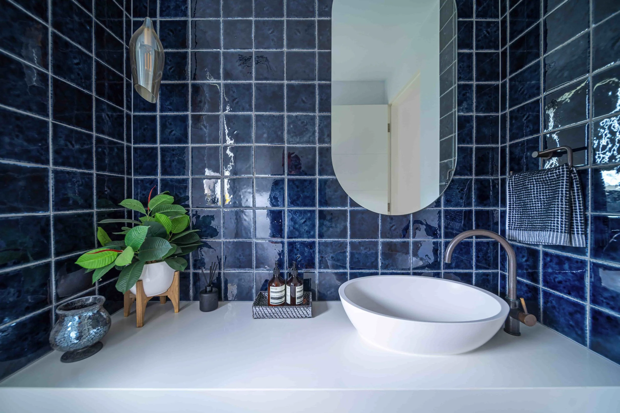 A modern bathroom with dark blue tiled walls, a white oval basin on bench, an oval mirror, a towel rack with a checkered towel, a potted plant on a small wooden stand, soap and lotion bottles, and a glass vase with a textured surface.