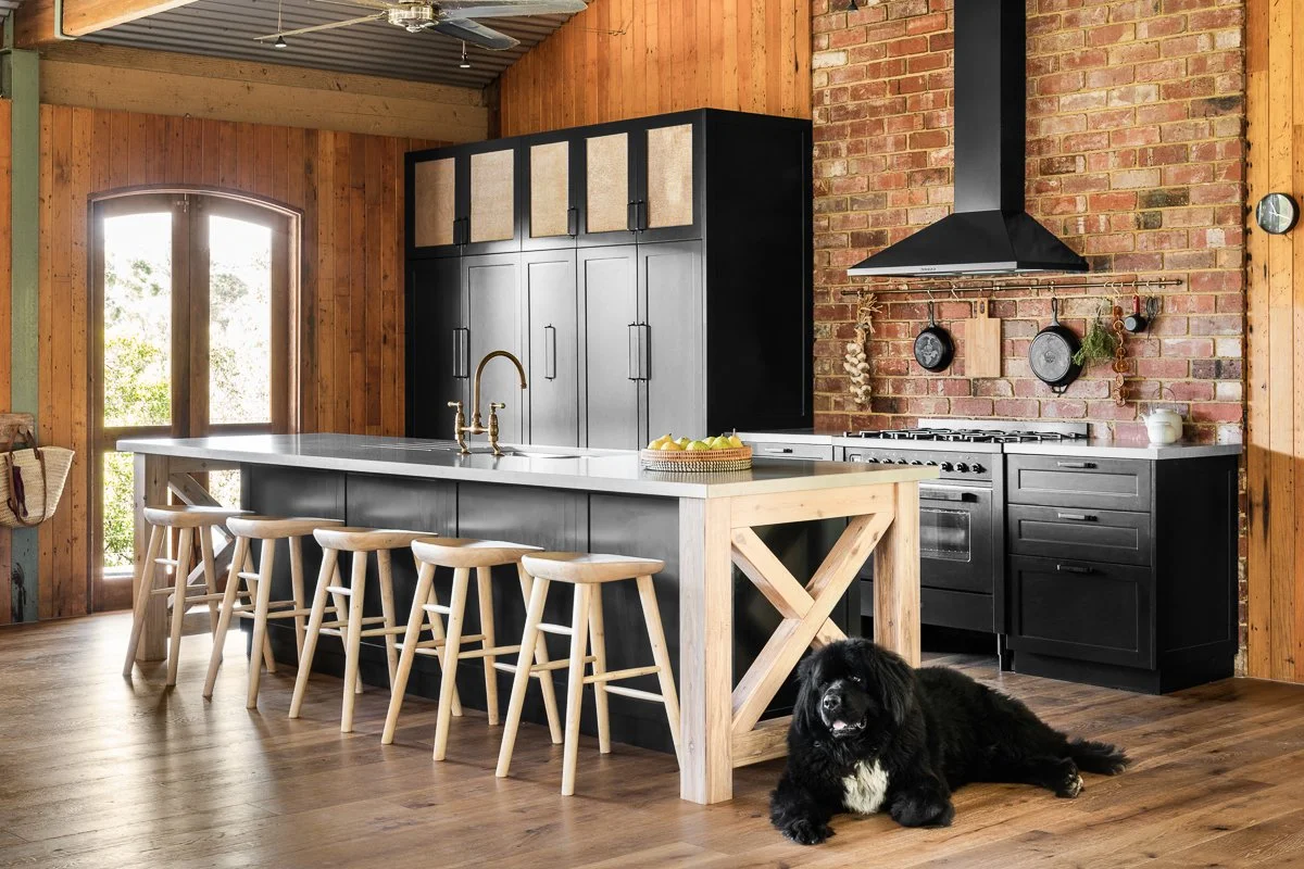A modern rustic kitchen with a large island, black cabinets, and a brick wall. A black dog is lying on the wooden floor near the island.