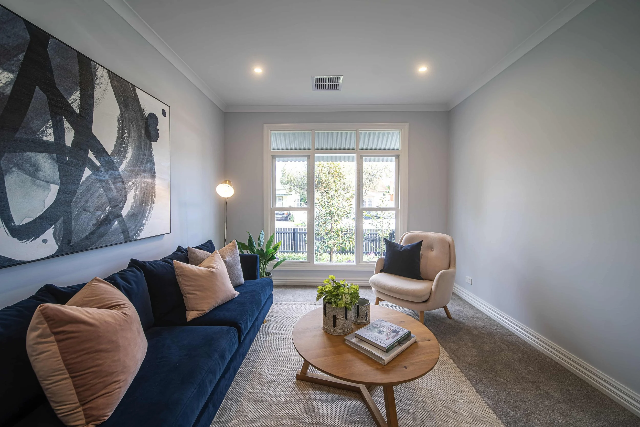 Formal living room or retreat area with a navy sofa, beige armchair, artwork on the wall, window, side table with plants, and books.