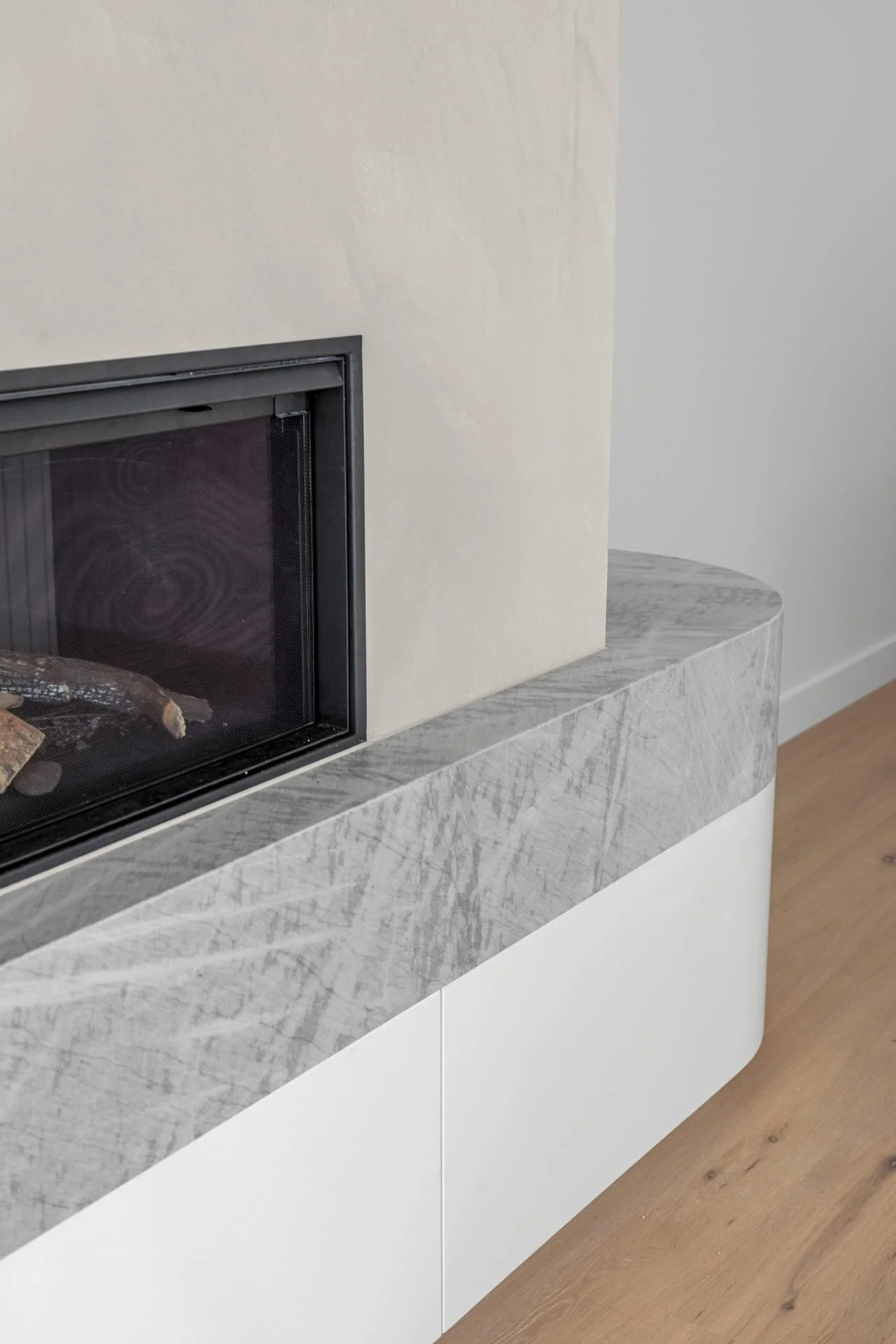 Close-up of a modern fireplace with a grey stone mantel and white cabinetry underneath, located in a contemporary living space with timber flooring.