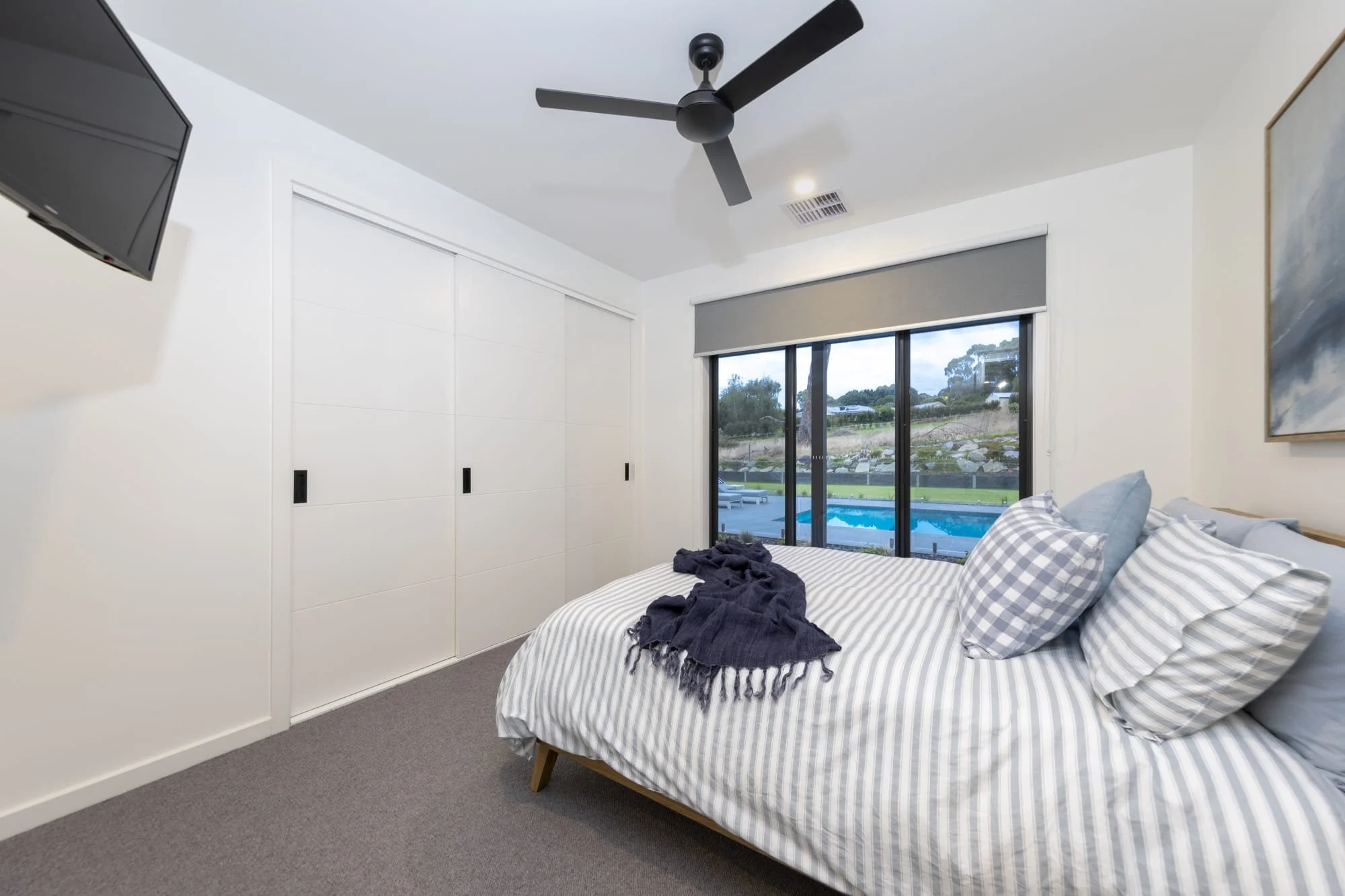 A bedroom with a bed covered in gray and white striped bedding and pillows, a window showing an outdoor pool and landscape, a closet with sliding doors, a wall-mounted TV, a ceiling fan, and a piece of wall art.