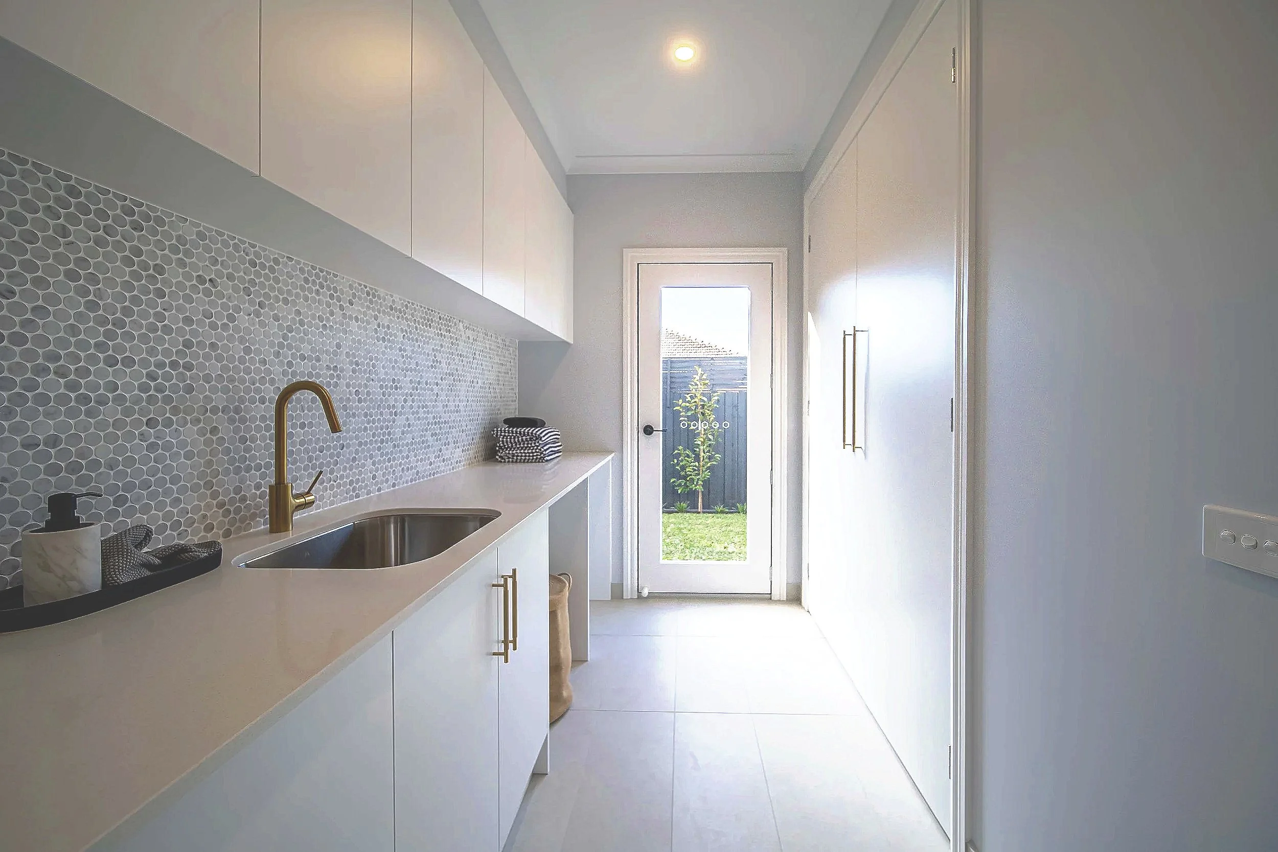 A bright, modern laundry with white cabinetry, a white stone benchtop, a round penny tile splashback, and a gold plated faucet and handles. There is a framed glass door at the end leading outside, with a small tree visible through the glass panel.