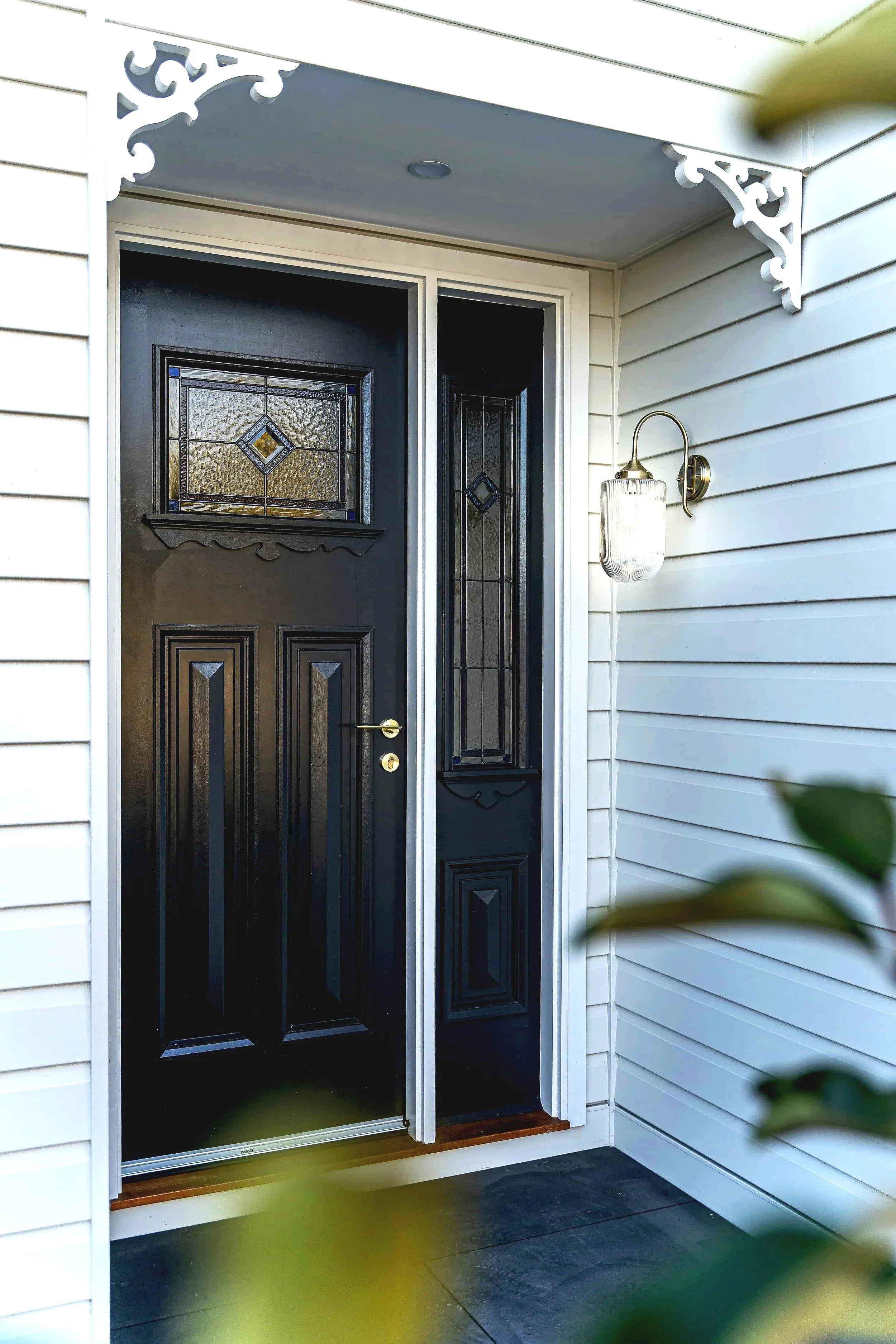 Front door of a house with black panel and decorative glass window, surrounded by white weatherboard walls and a porch light, partially obscured by some out-of-focus green leaves in the foreground.