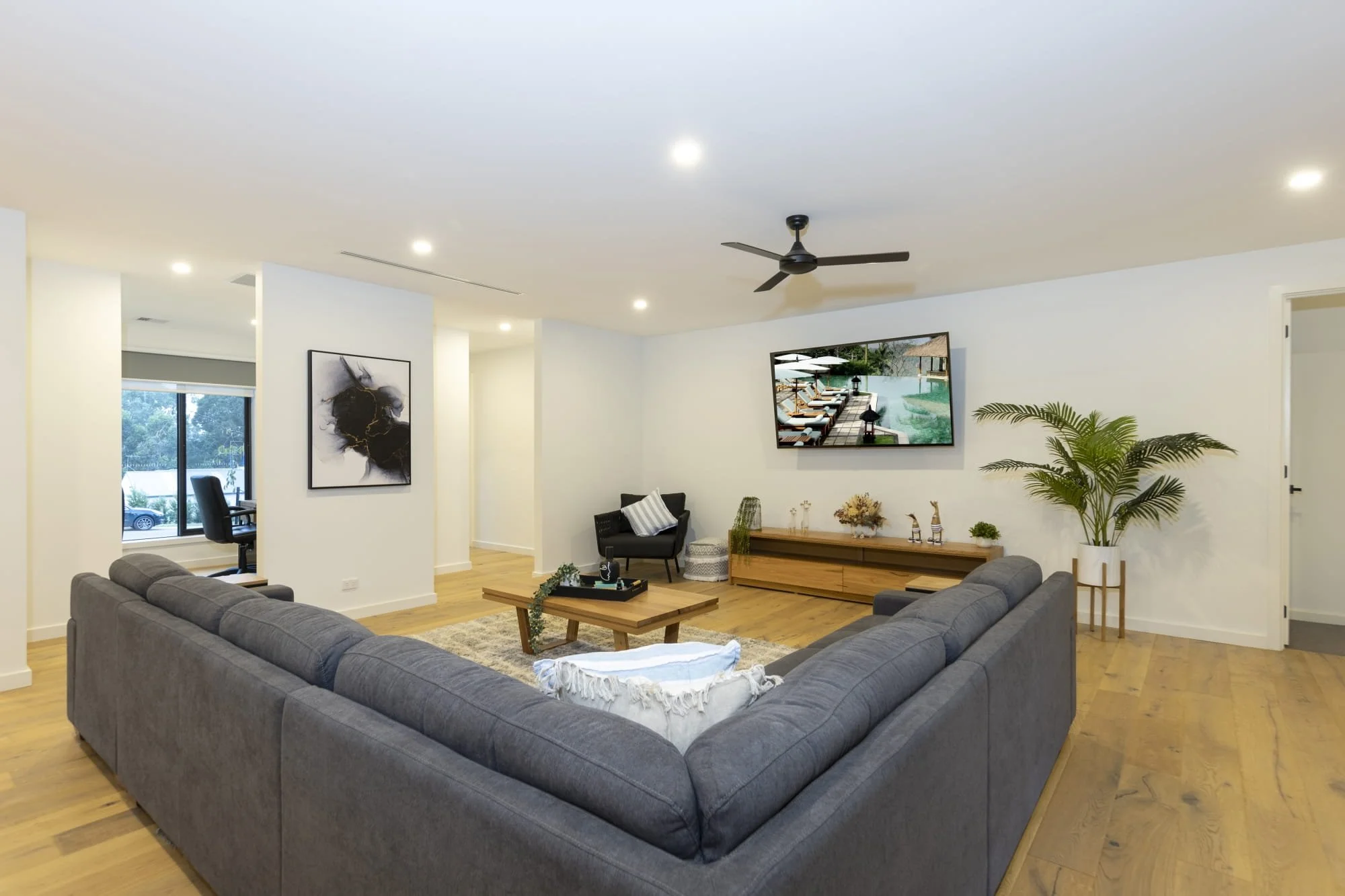 Contemporary living room with a large grey sectional sofa, wooden coffee table, flat-screen TV, artwork on the wall, potted plant, and hardwood flooring.