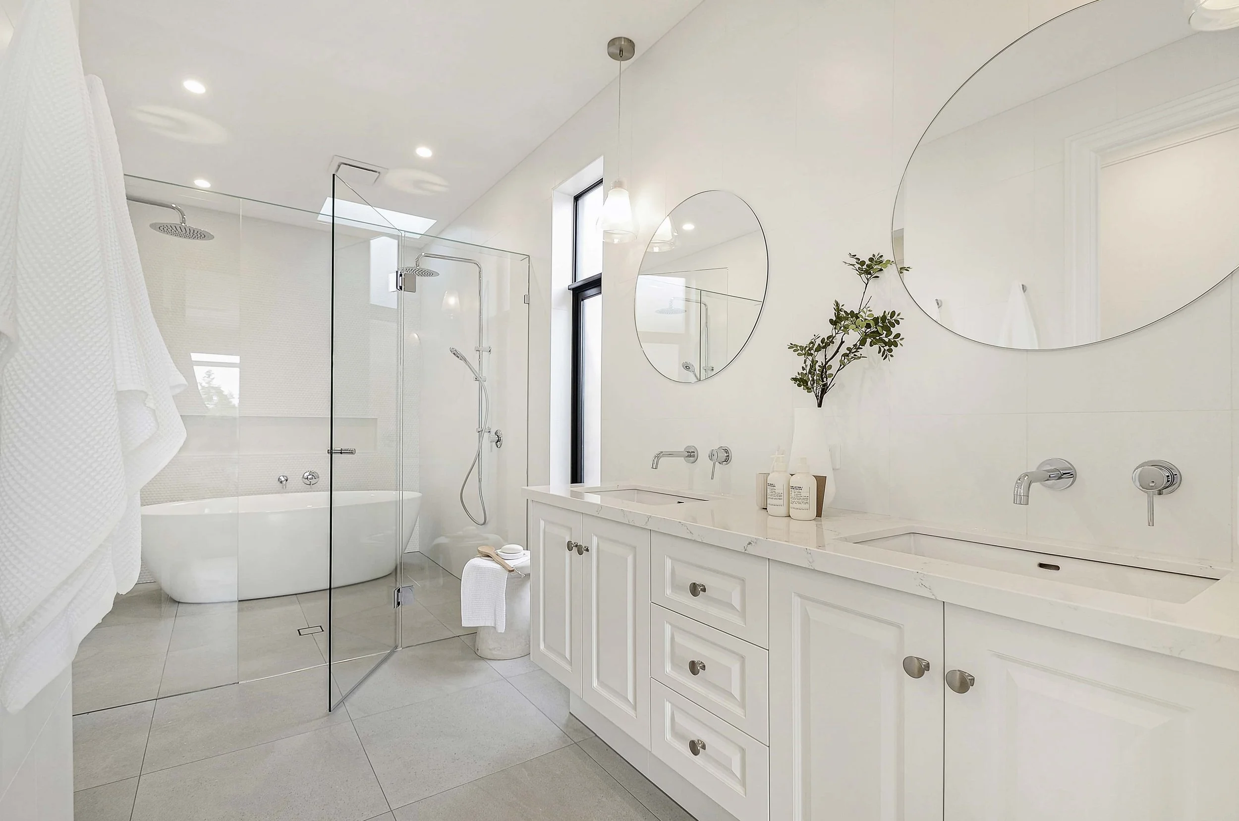 Modern white ensuite with double basin vanity, round mirrors, and a glass-enclosed wet area. The wet area contained two shower rails and an oval white bathtub. Natural light streams in from the window, and the decor is minimalist.