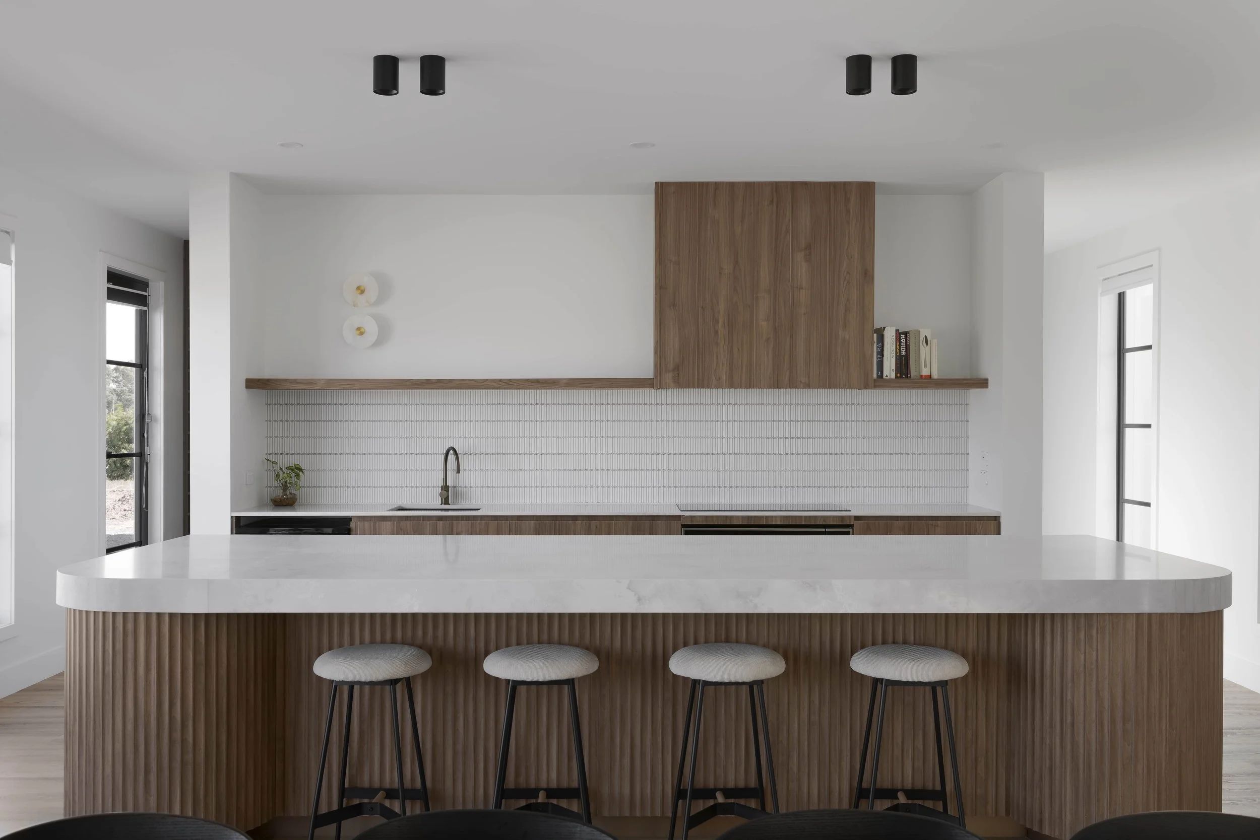 Modern kitchen with thick white stone benchtop, timber cabinetry with a scalloped timber island, black stools, and large windows. The back kitchen wall is minimal with a boxed timber rangehood, two circular wall sconces and a floating timber shelf.