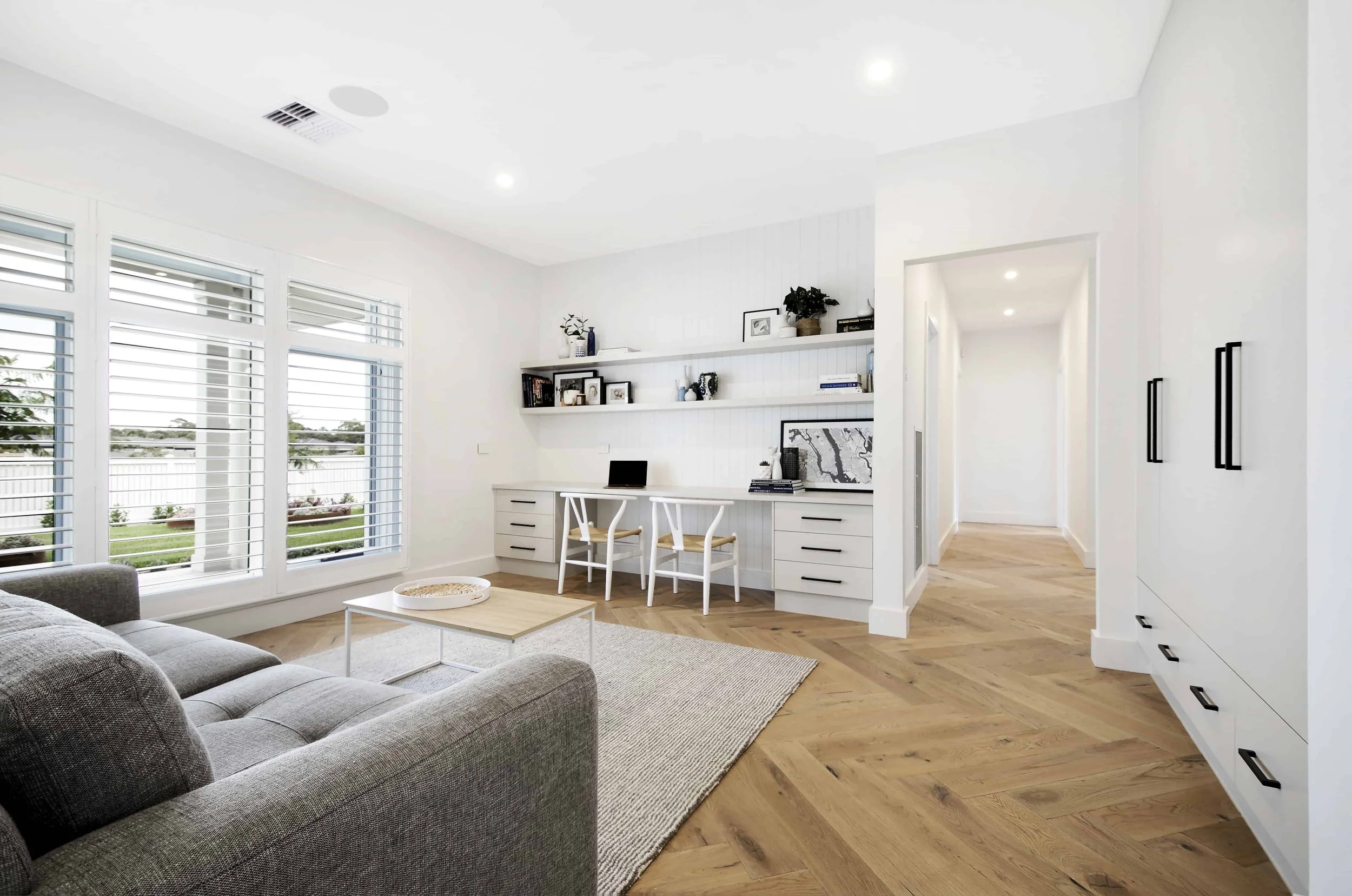 Private retreat lounge and study with in built desk cabinetry and wardrobes featuring black handles. The recessed wall behind the study area is lined in VJ board and has two white floating shelves spanning the full length.