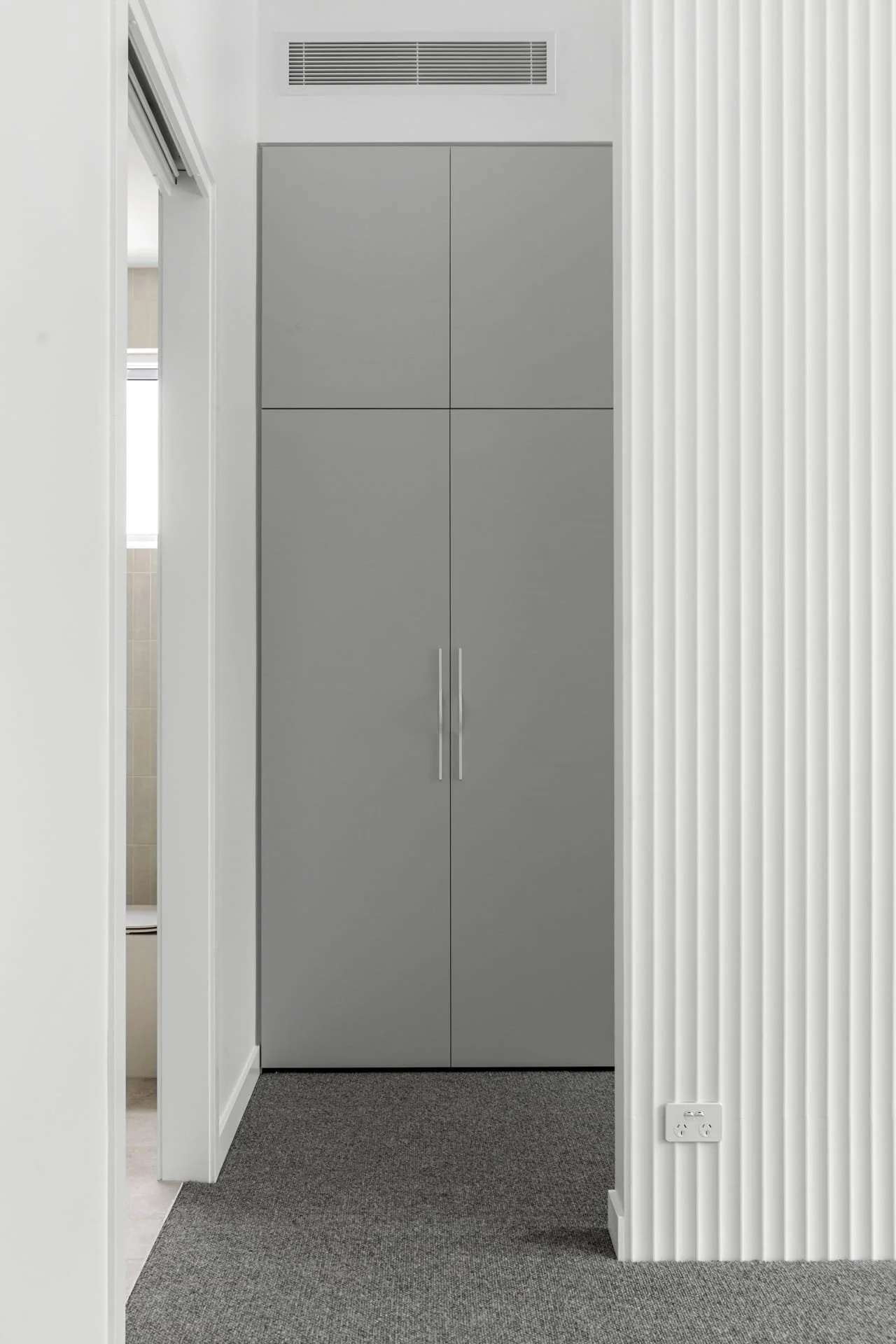 Grey double door wardrobe and chrome handles, in a room with white walls and white scalloped wall panelling. Floor is carpeted, and there are electrical outlets near the baseboard.