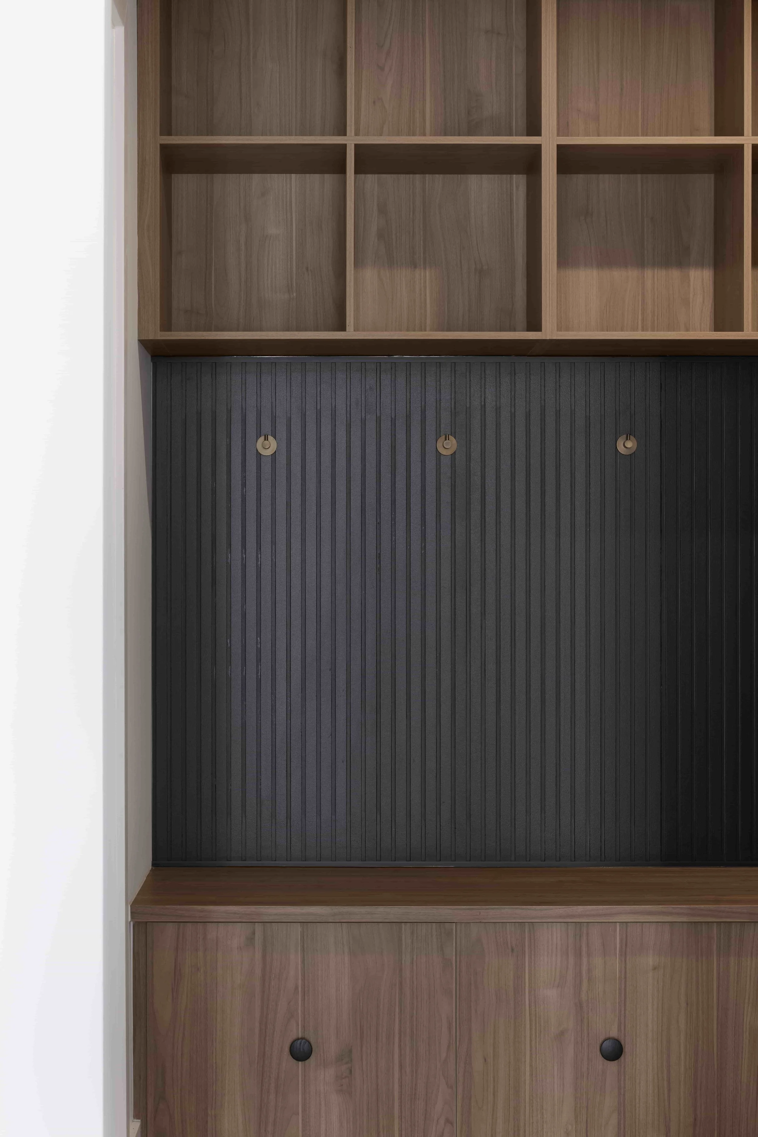 A built-in wooden cabinet in a mud room with a black vertical back panel and metal hooks. The upper part features open pigeon hole shelving in a natural wood finish, and the bottom has closed storage with black knobs.