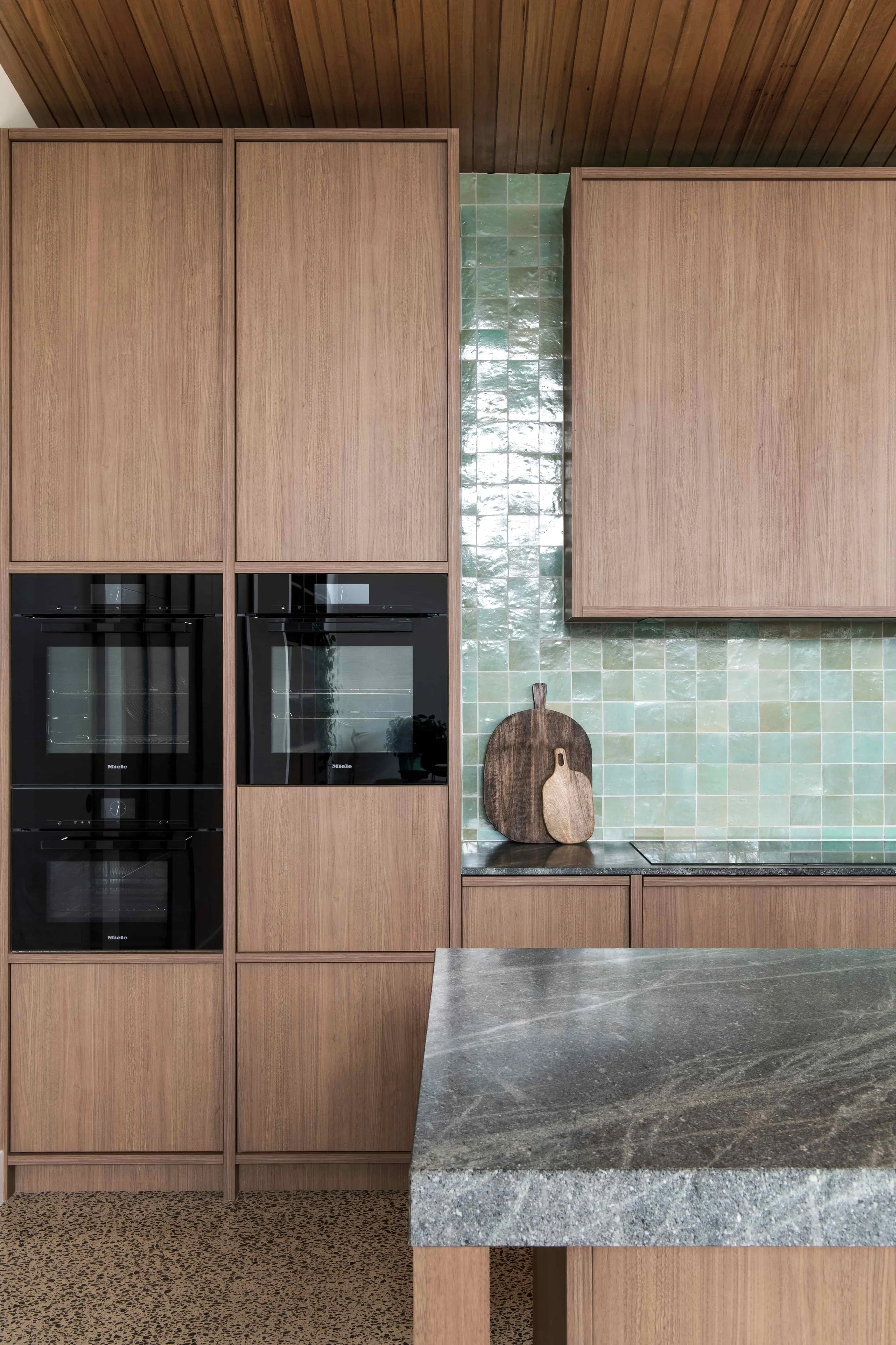 Kitchen with timber cabinetry, two built-in black ovens, green mosaic tiled wall, and a grey stone benchtop.