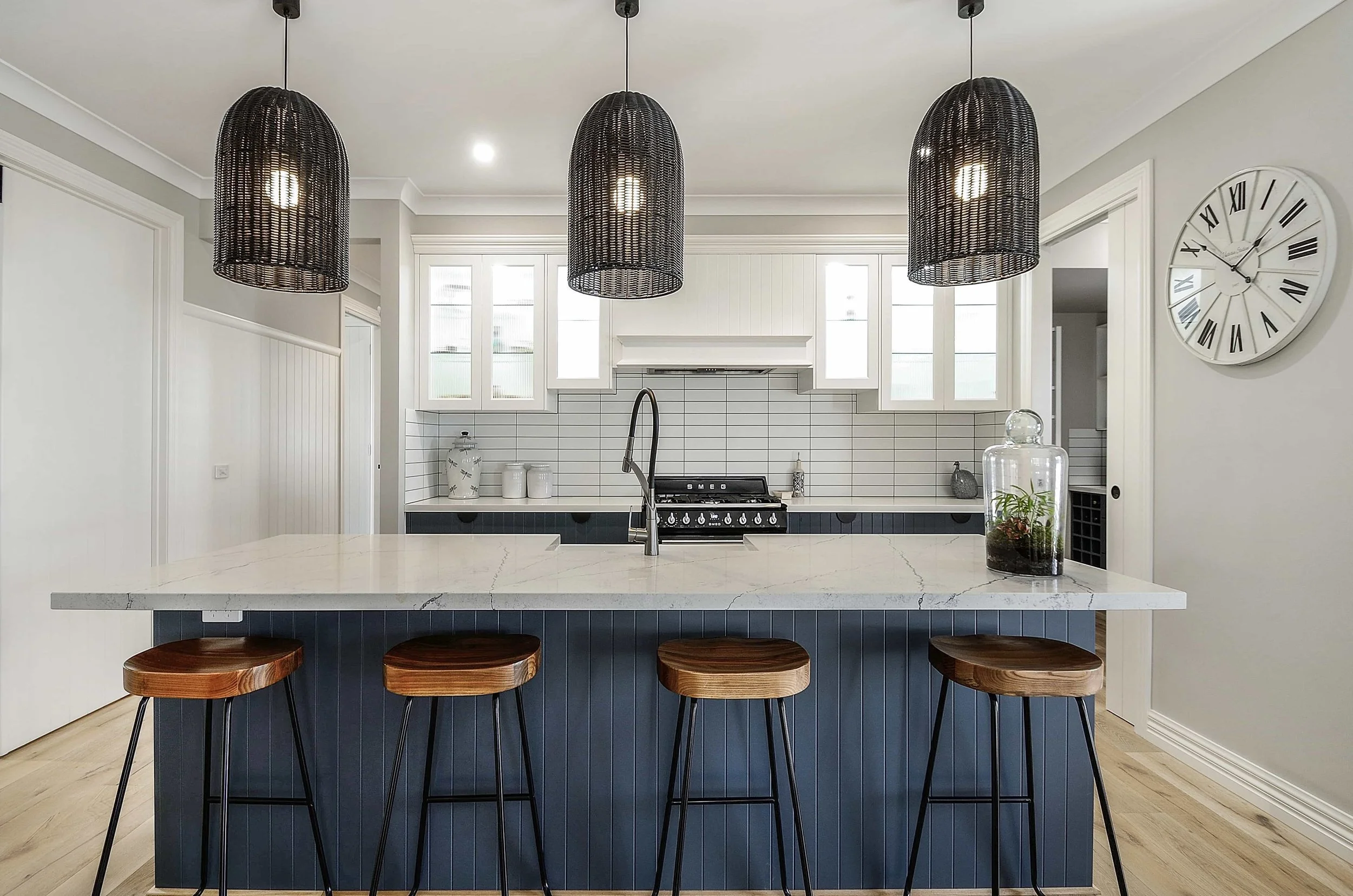 Modern kitchen with a blue island, white stone benchtops, black rattan pendant lights above the island, and bar stools with wooden seats. Light-coloured wood flooring and grey walls.. The island has vertical routing in the panels.
