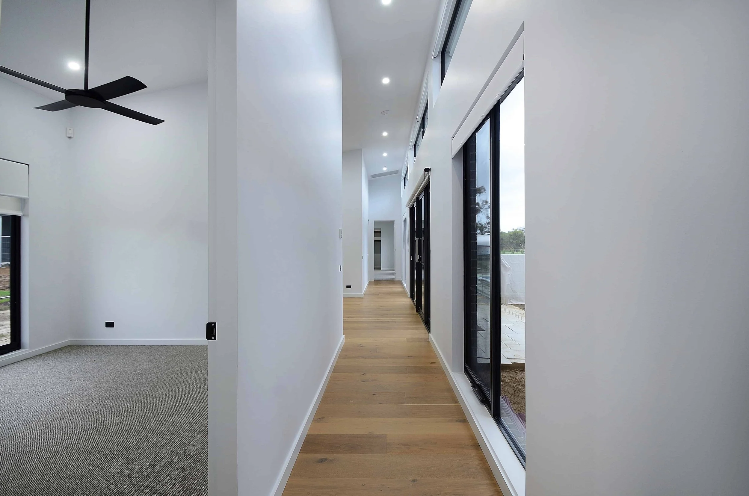 A modern interior hallway with high white walls, large black-framed glass doors and windows, wood flooring, and ceiling lights. A ceiling fan hangs from the white ceiling. The ceiling is raked.