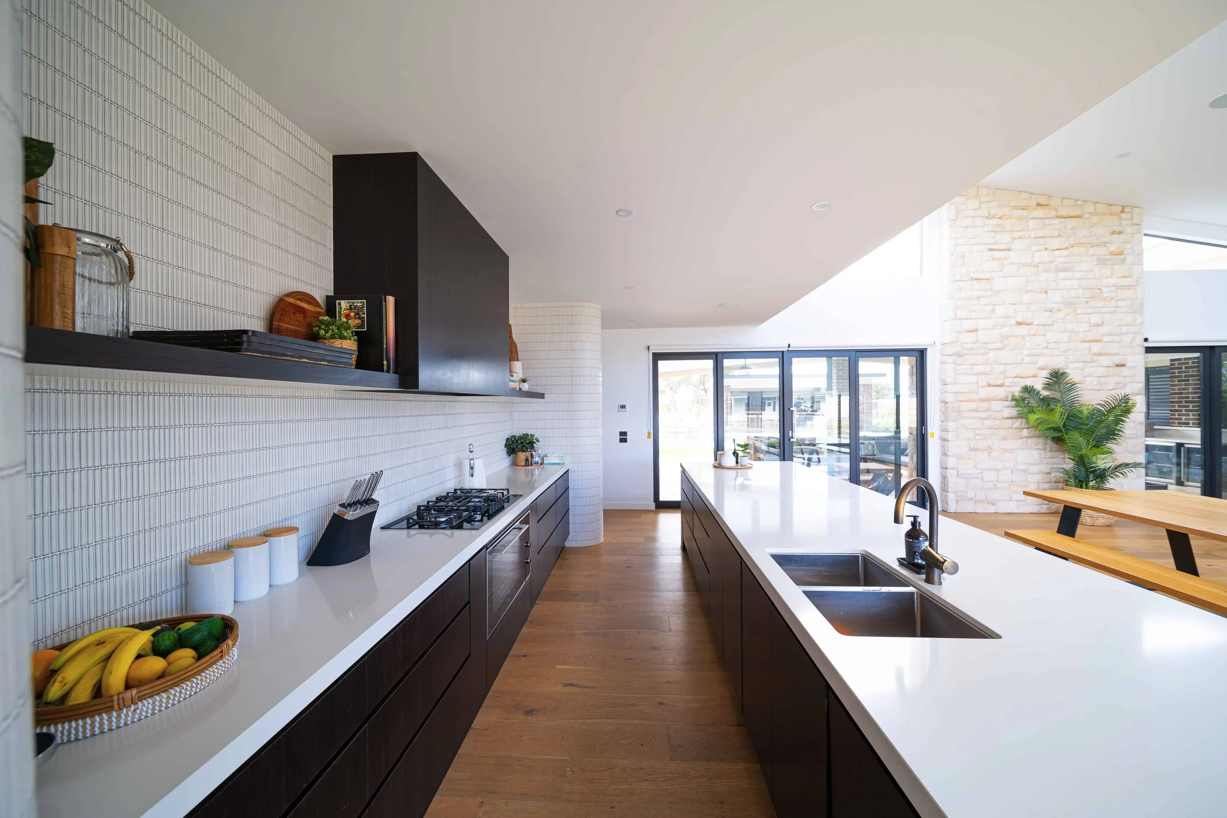 Modern kitchen with a white stone benchtop, black cabinets, and large windows in the background. The kitchen has a floating shelf either side of the concealed rangehood. There is a curved tiled accent wall on either side of the back kitchen bench.