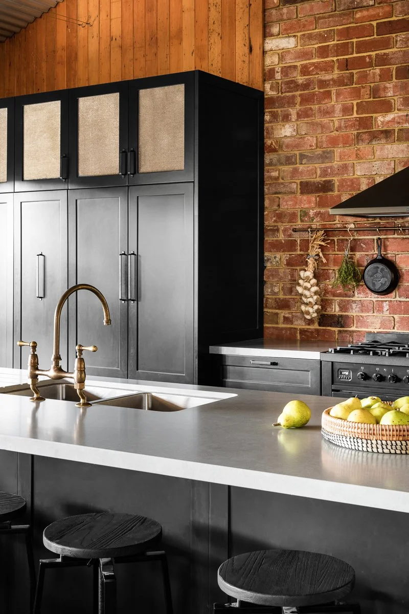 A modern rustic kitchen with black cabinets, brick wall, and antique copper faucet on grey stone benchtop with a bowl of pears.