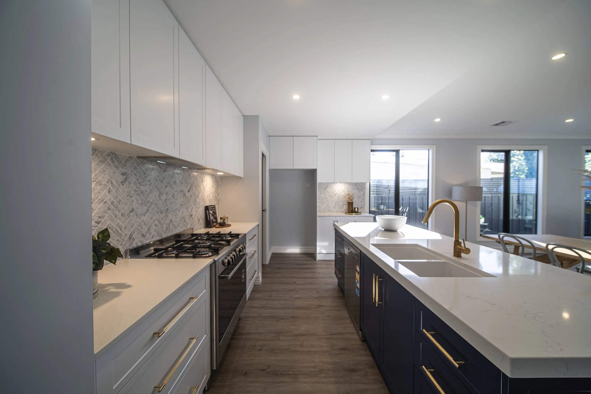 Modern kitchen with white and navy cabinets, grey herringbone pattern splashback, and a large island with a white benchtop. The faucet and handles are gold plated.