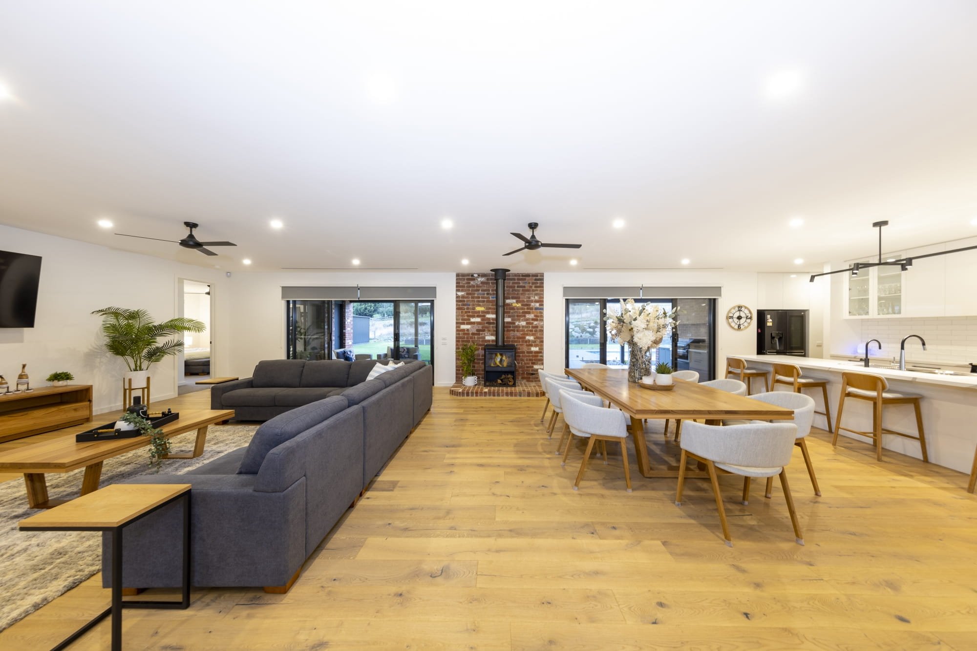 Spacious, modern living room with white walls, hardwood floors, a brick fireplace, ceiling fans, large sliding doors, a grey sectional sofa, a wooden coffee table, plants, and a dining area with a wooden table and white chairs.