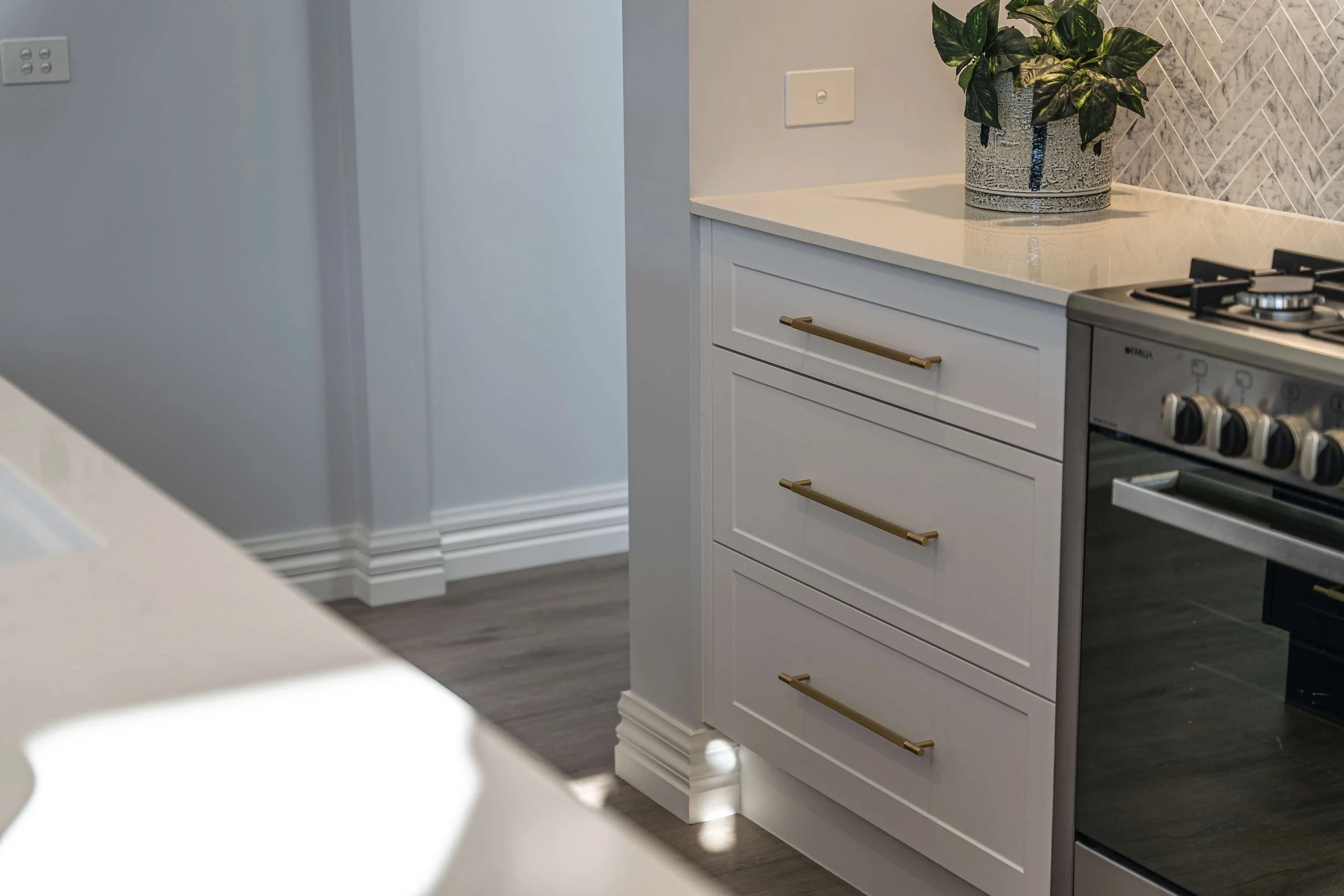A section of a modern kitchen with white cabinets featuring gold handles and a benchtop with a potted plant. A stainless steel stove is partially visible. The wall has a herringbone tile pattern.
