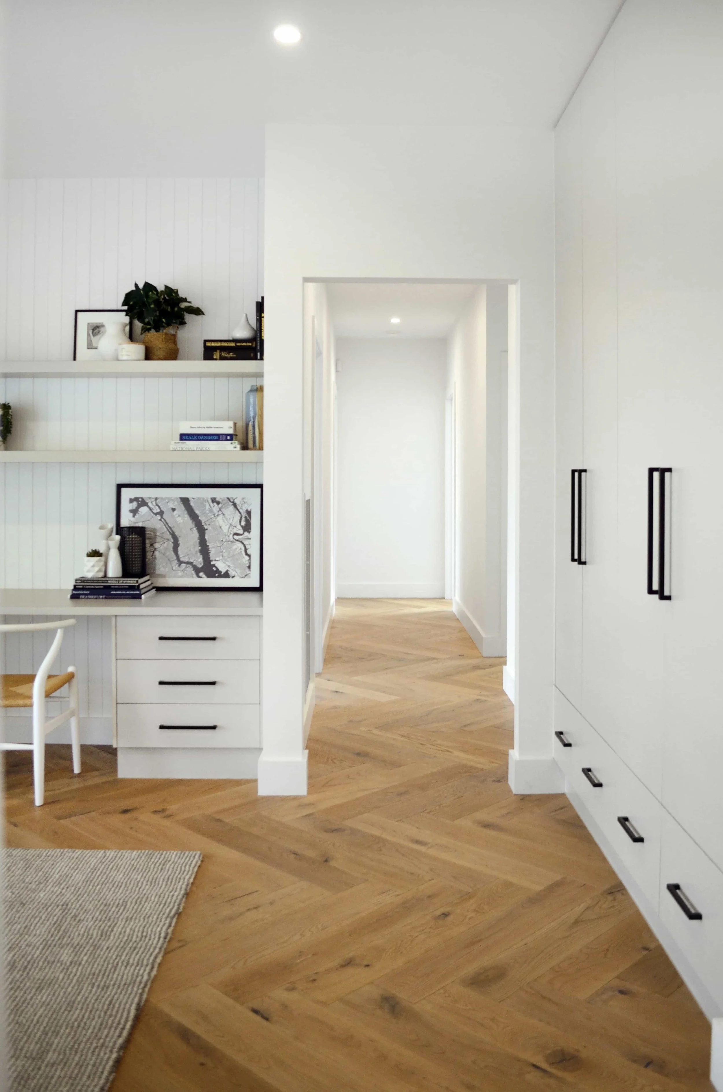 View of a study to a hallway with built in desk cabinetry and wardrobes featuring black handles. The recessed wall behind the study area is lined in VJ board and has two white floating shelves spanning the full length.