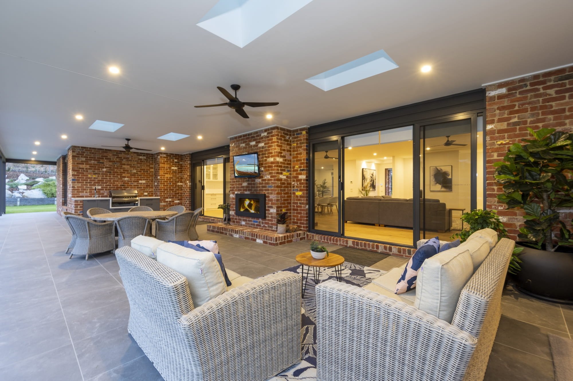 Covered outdoor alfresco with wicker chairs, a small round table, a red brick fireplace and hearth, ceiling fans, and skylights, adjacent to a house with large sliding glass doors and a view of the interior living space.