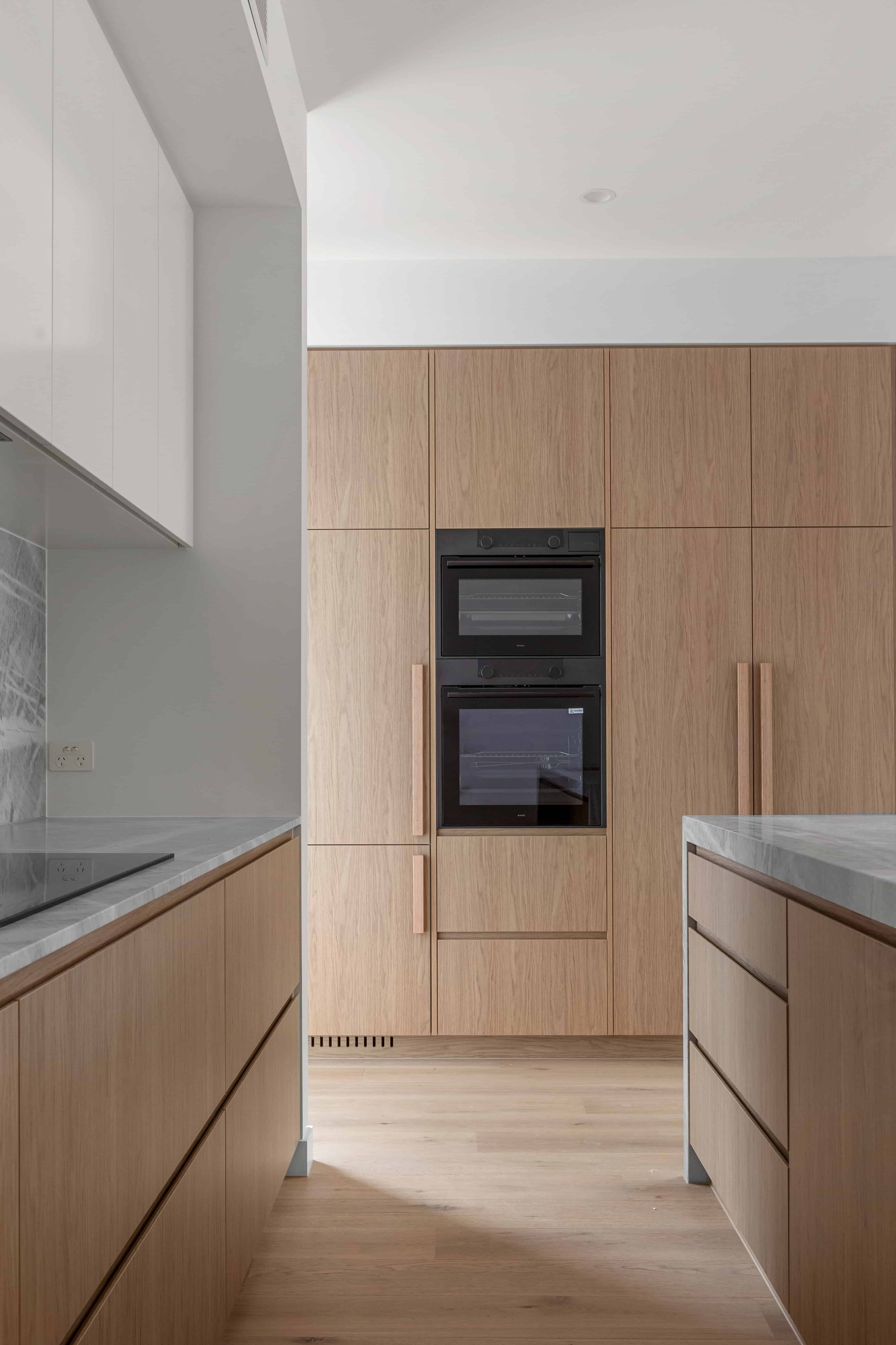 Modern kitchen with light timber cabinets, built-in black ovens, grey stone benchtops, and light timber flooring.