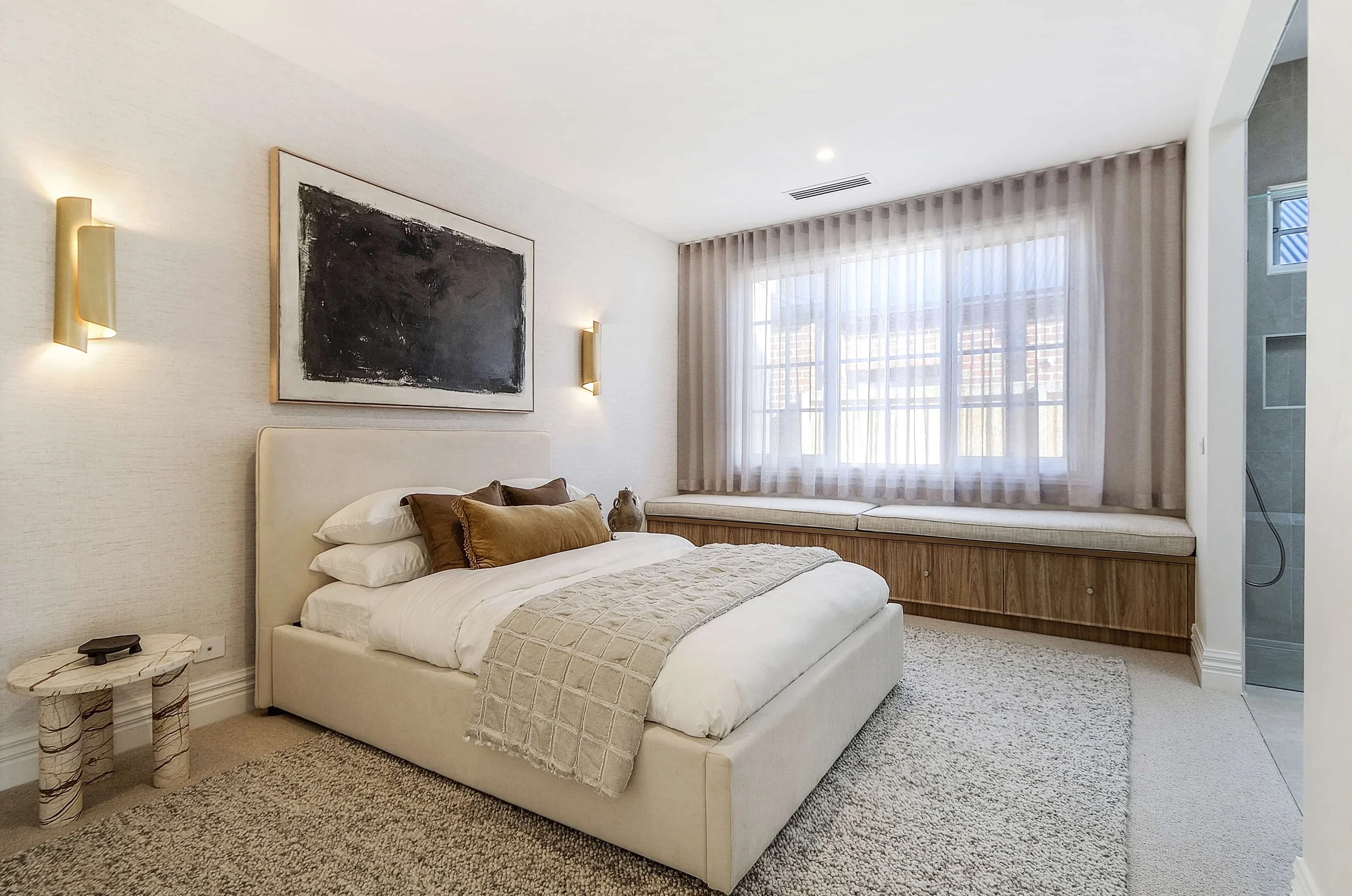 A modern master bed suite with wall-mounted feature lights, abstract artwork, a large window with sheer curtains, and a built-in window seat with storage underneath. The overall decor is minimalist and neutral.