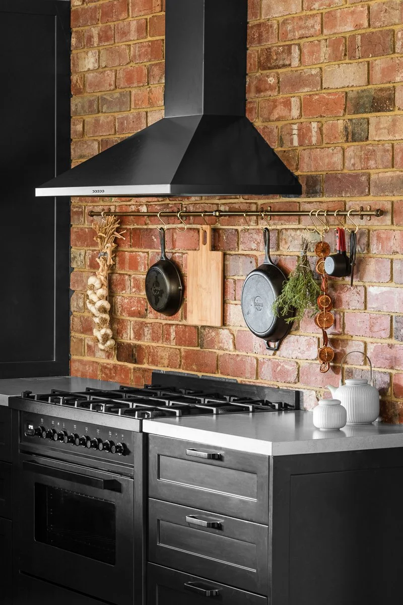 A modern rustic kitchen with a brick wall, black cabinets, a black range hood, and hanging pots and herbs on a brass rod.